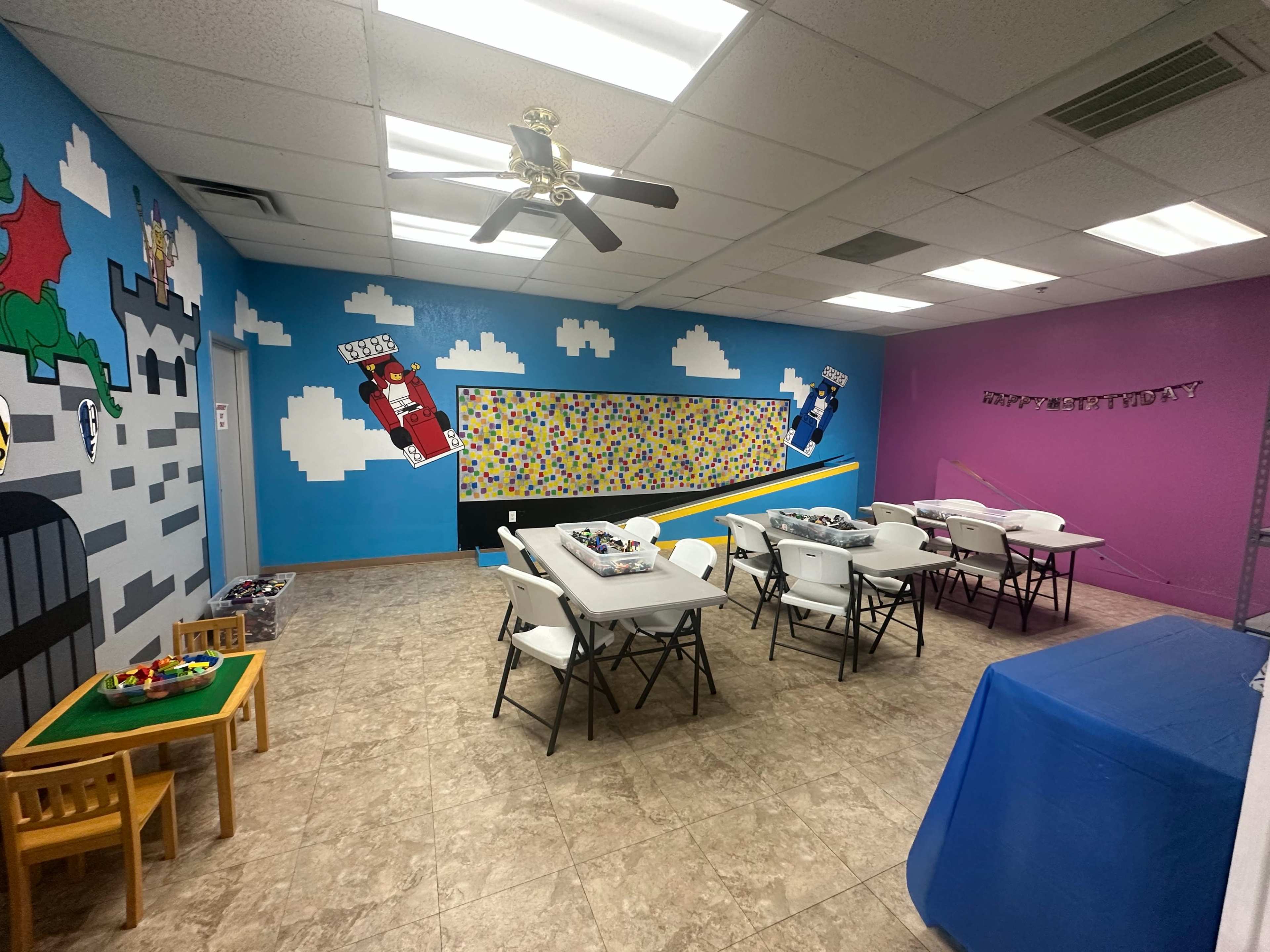 The image shows a brightly colored classroom with tables and chairs arranged for activities, featuring murals of superheroes and a bulletin board.