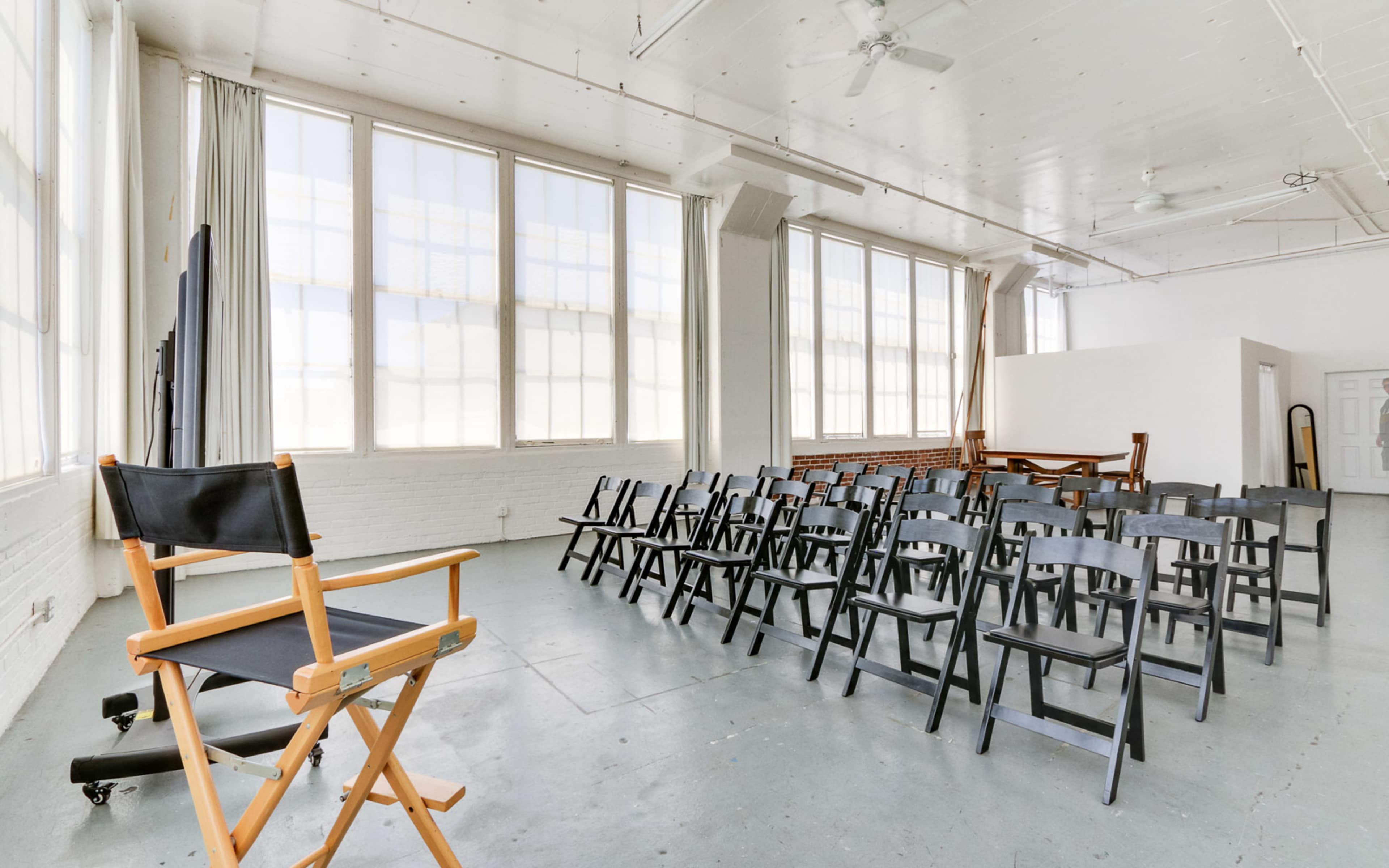 A bright, spacious room with large windows features rows of black chairs facing a wooden director's chair.