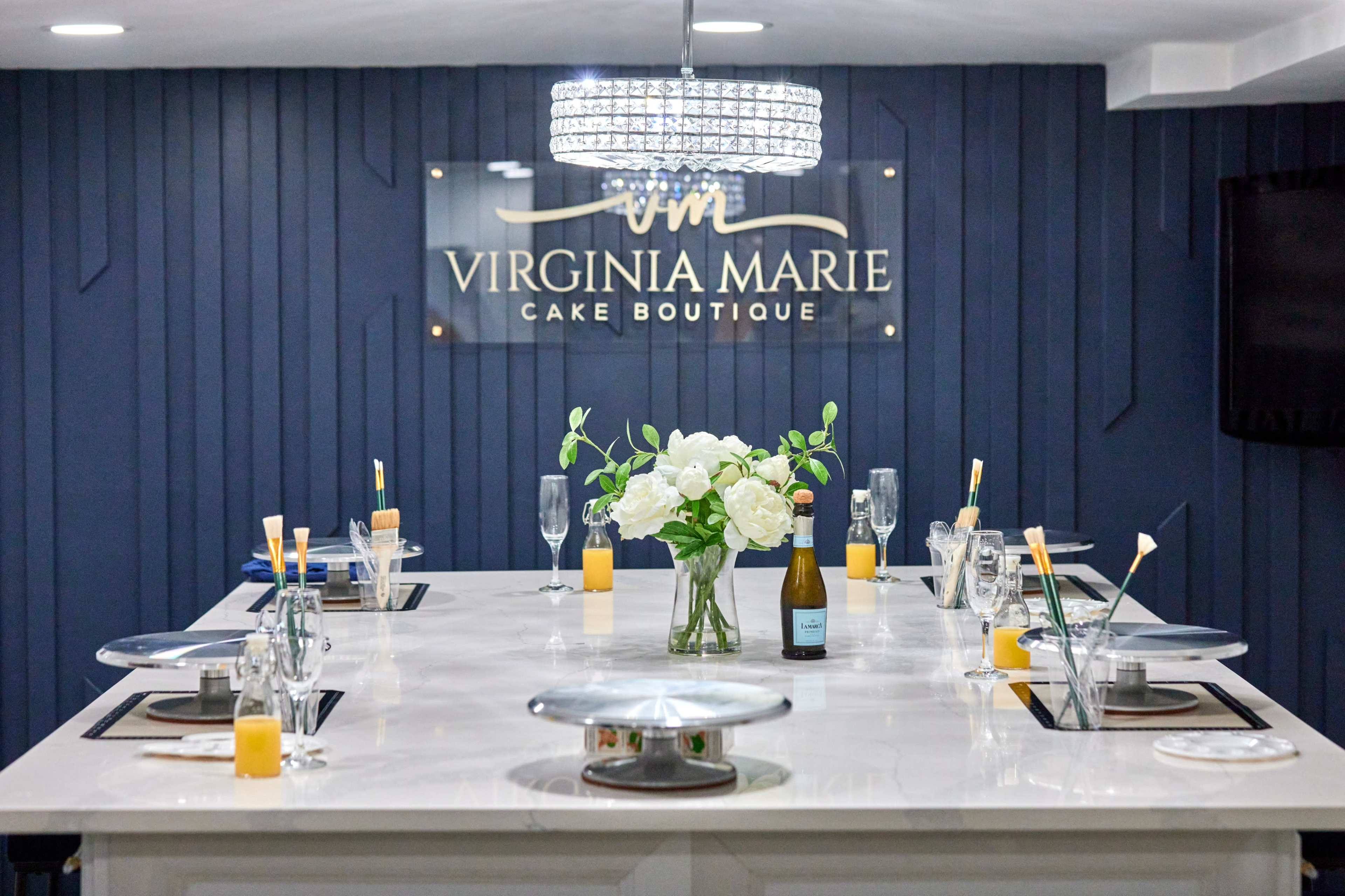 The image shows a modern cake boutique with a large, elegant table set for a gathering, featuring a floral centerpiece, sparkling chandelier, and branded wall sign.
