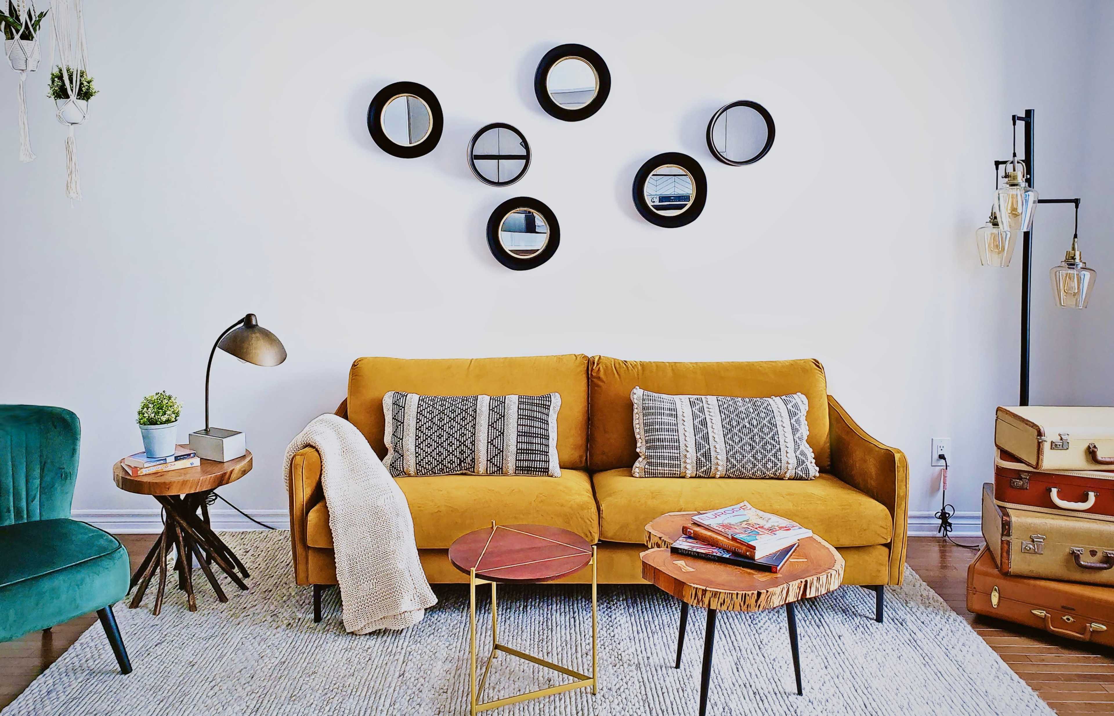 A cozy living room featuring a mustard yellow sofa with patterned cushions, surrounded by two side tables, a floor lamp, and a wall adorned with circular mirrors.