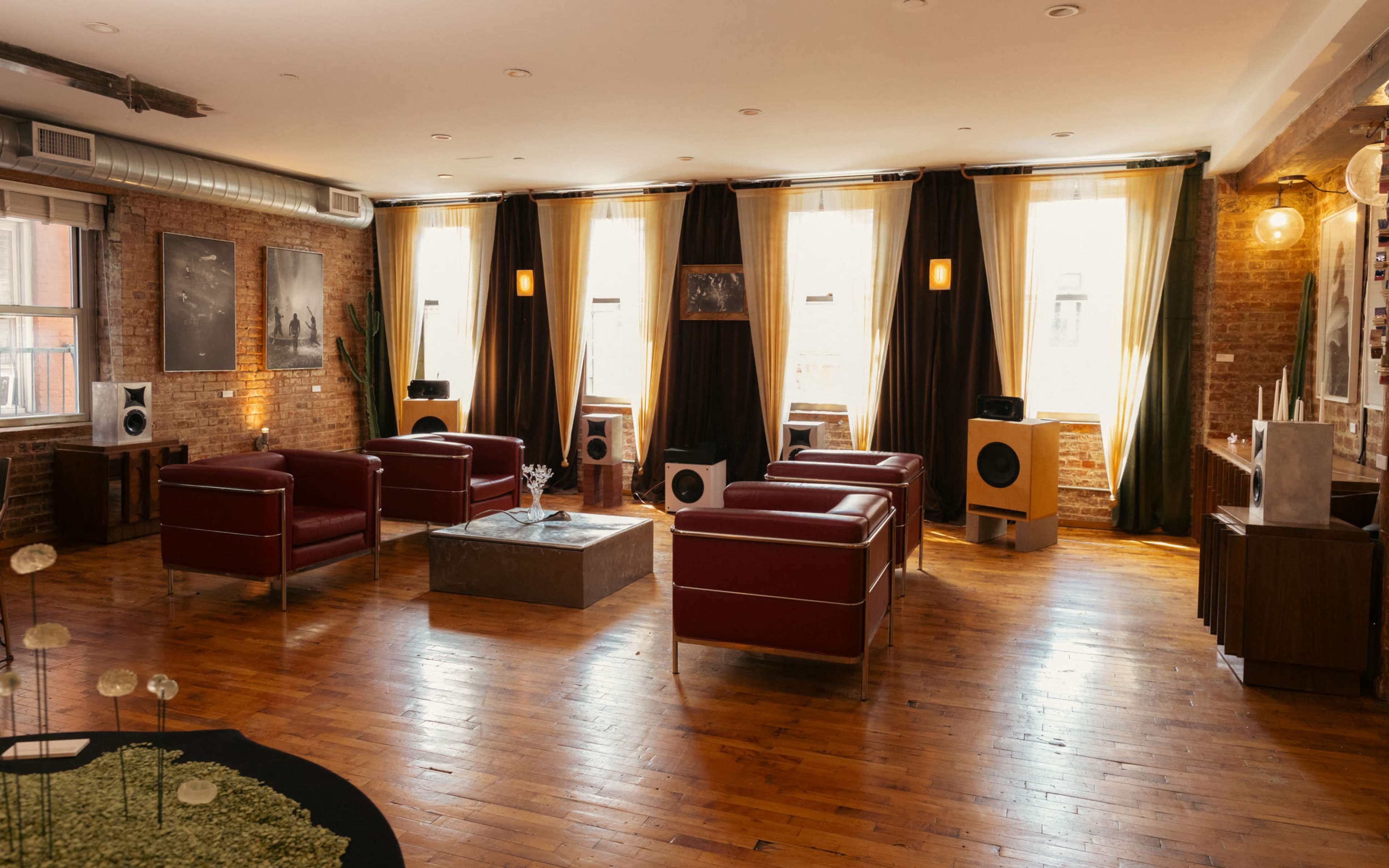 The image shows a spacious room with hardwood floors, featuring red leather chairs, large windows with curtains, and various decorative elements.