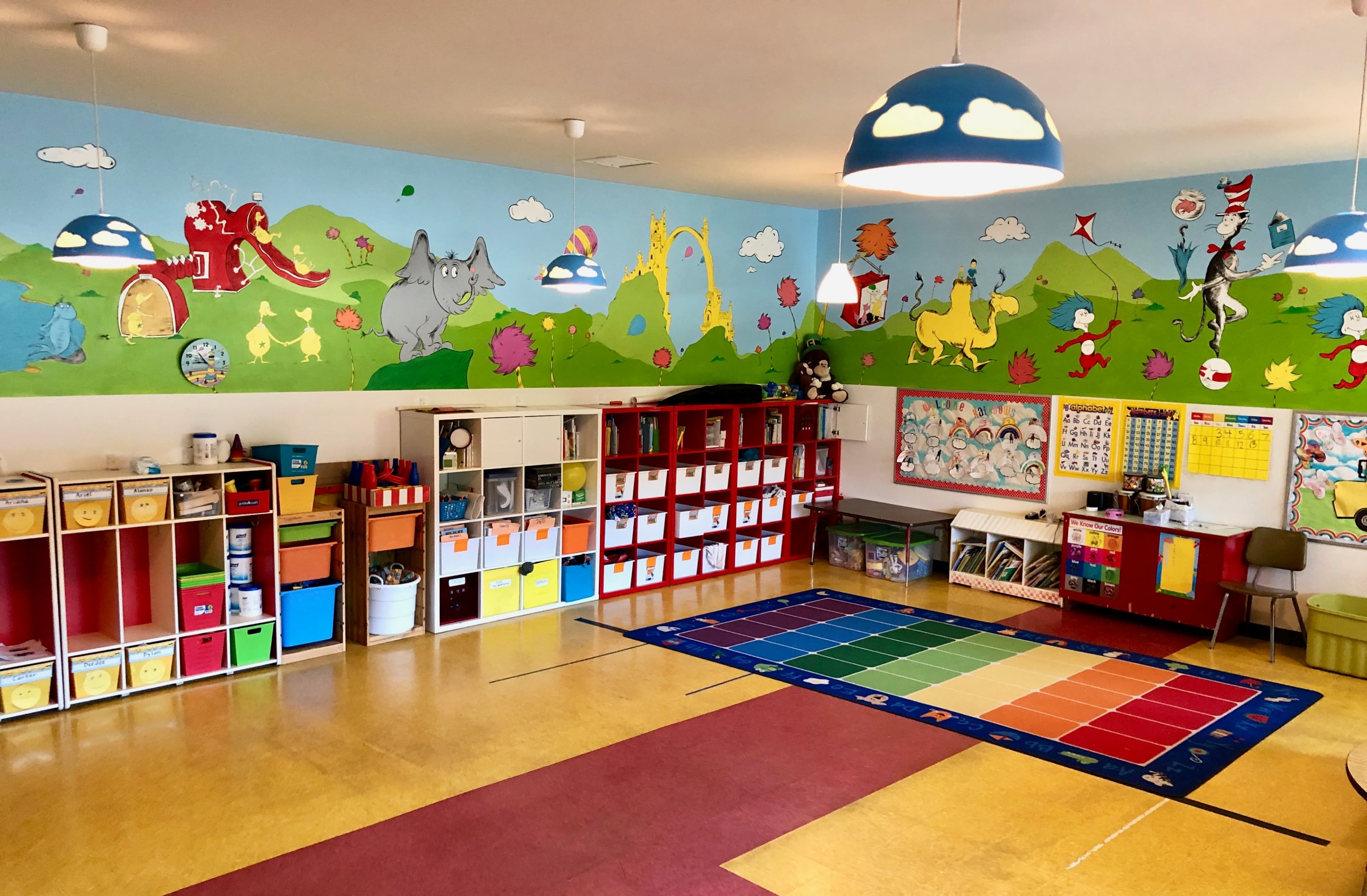 A brightly decorated kindergarten classroom featuring colorful wall murals, storage units, a play area with a multicolored rug, and various educational materials arranged on the walls.