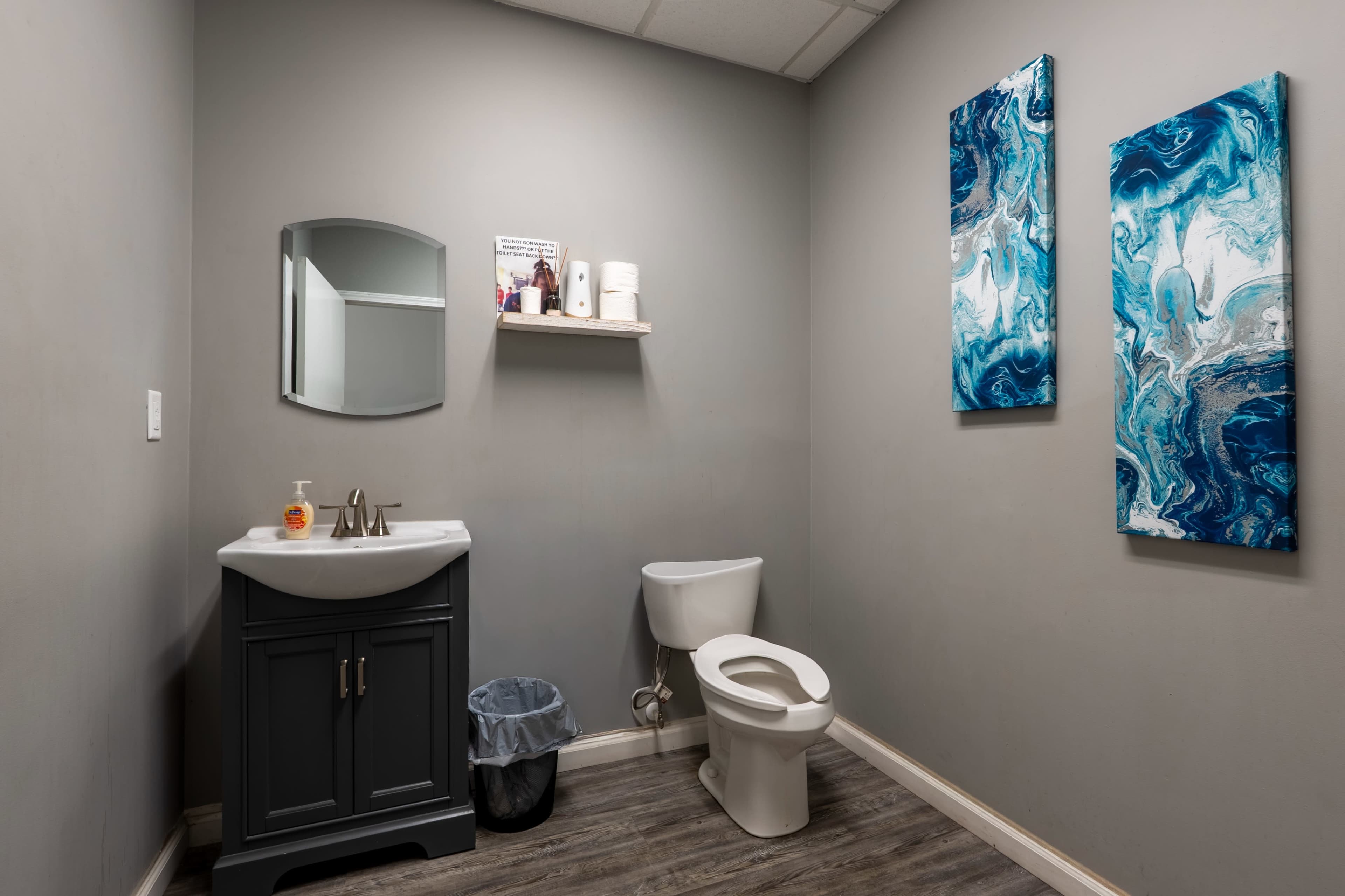 The image shows a small bathroom with a dark cabinet sink, a toilet, a mirror, and two abstract blue and white artworks on the wall.
