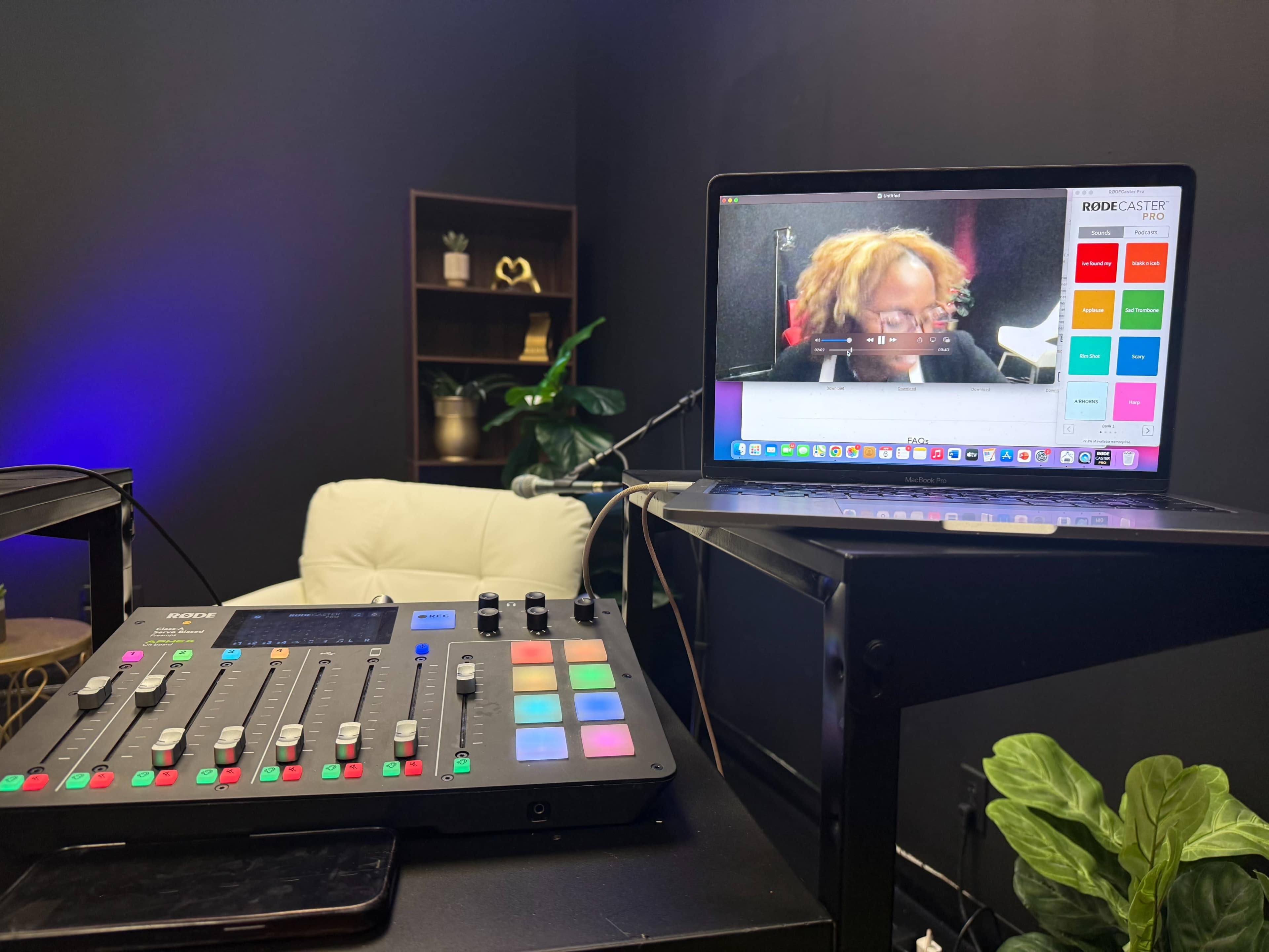 A digital audio mixer is positioned in the foreground, while a laptop displays a video call with a person, set against a dark backdrop featuring a shelf and plants.