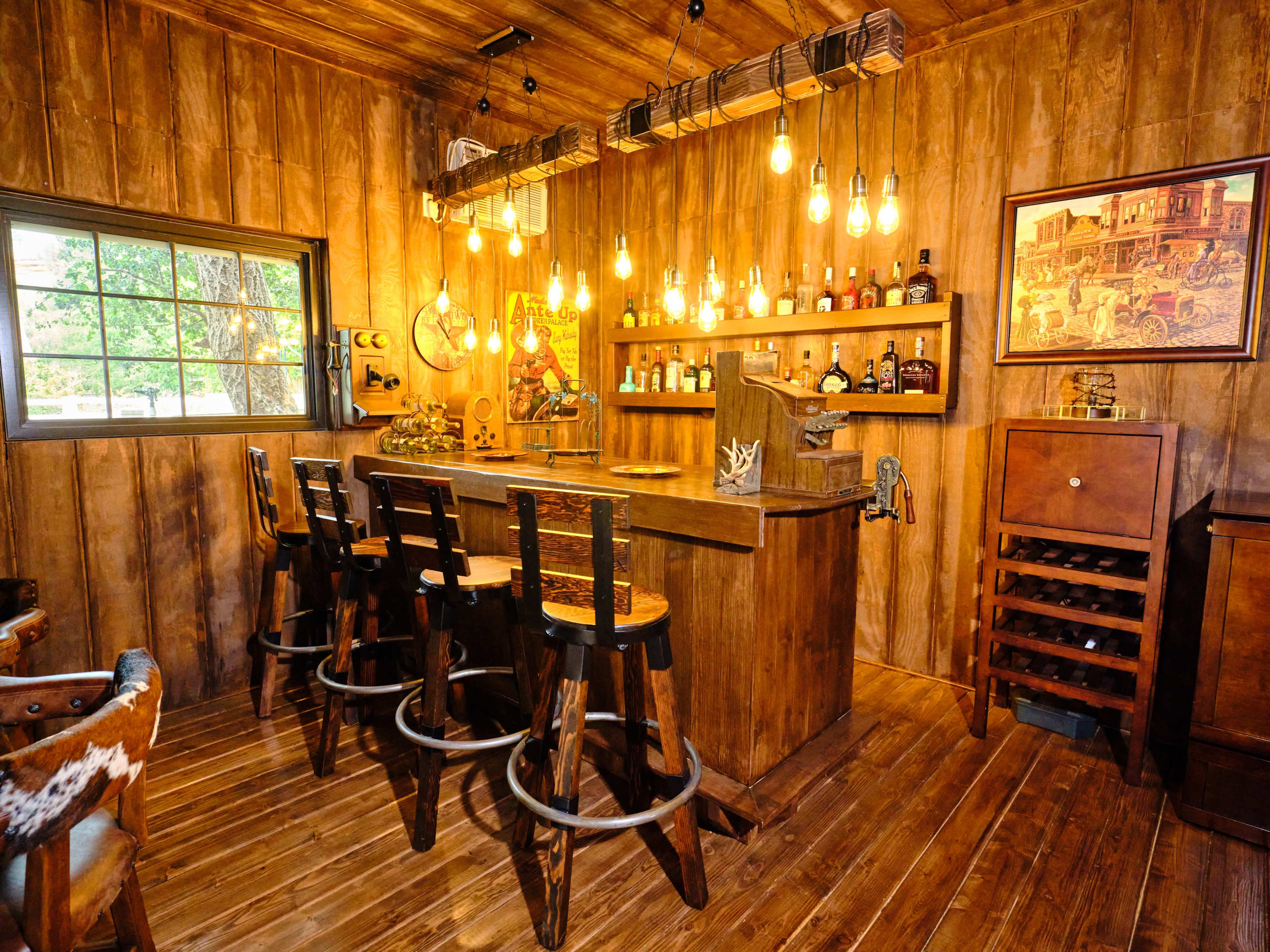 A rustic wooden bar area with four stools, a variety of bottles displayed on a shelf, and warm lighting fixtures hanging from the ceiling.