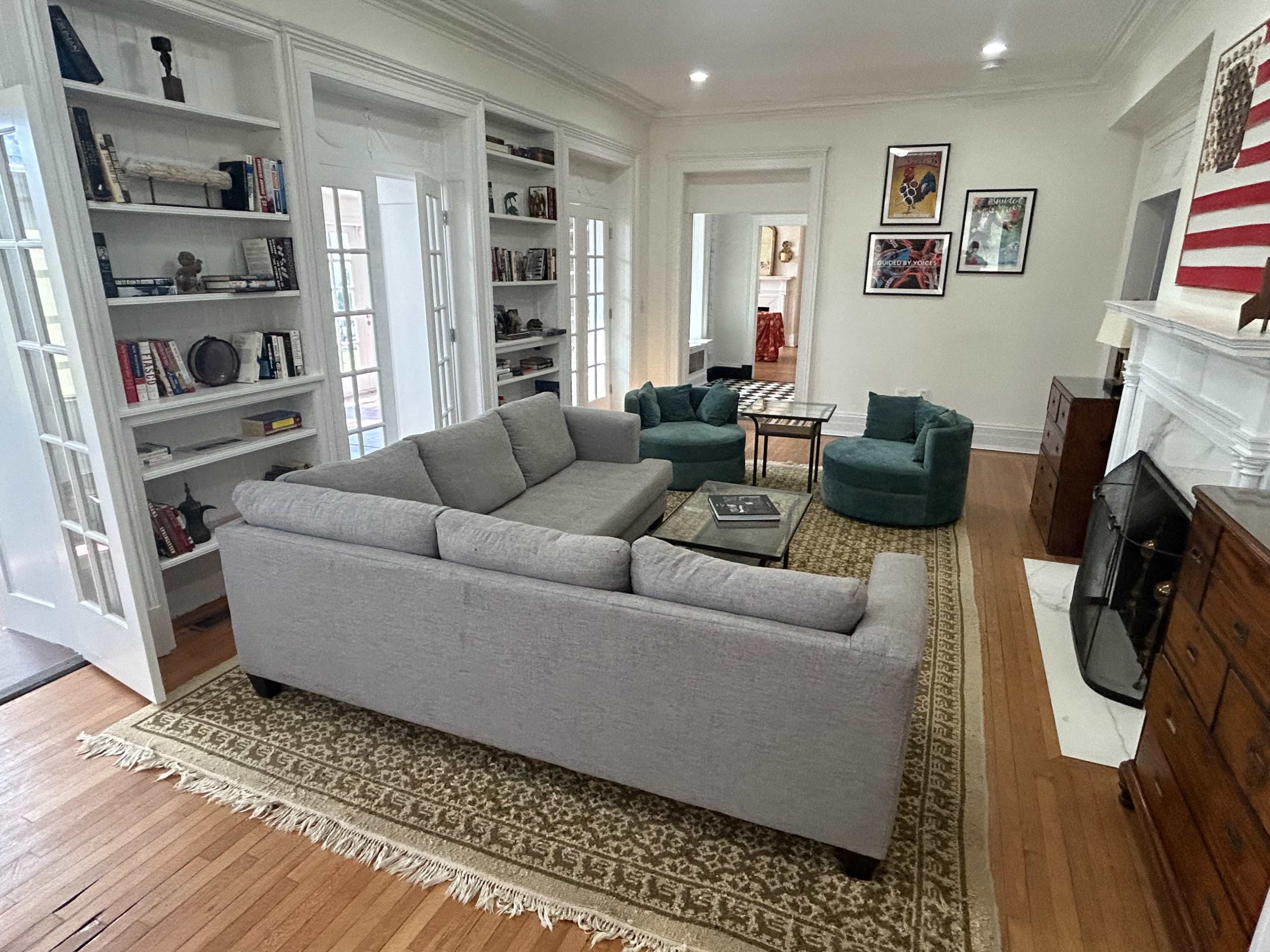The image depicts a living room with a gray sofa, two green armchairs, a coffee table, bookshelves filled with books, and a fireplace on a wooden floor.