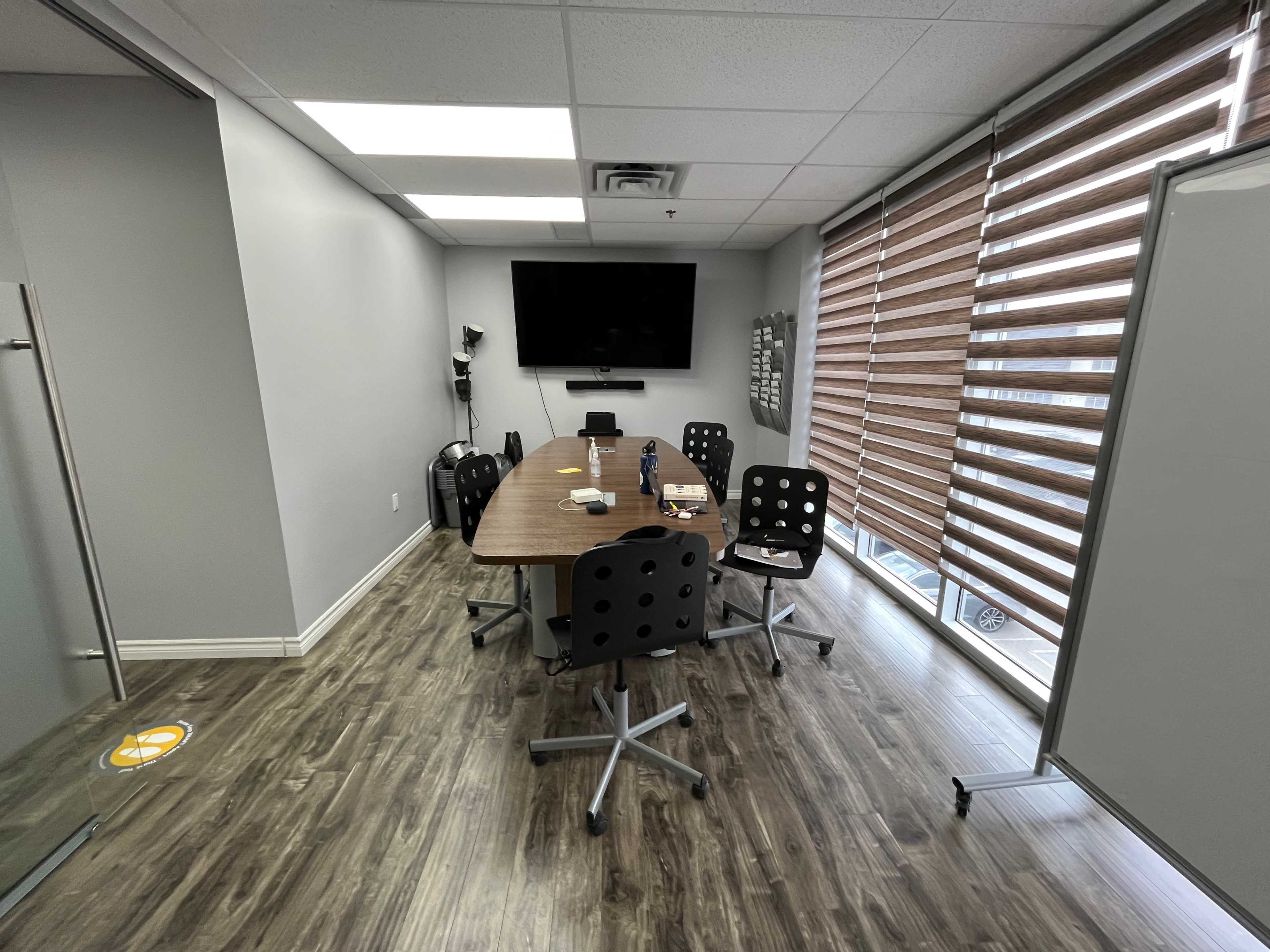 A modern conference room features a long wooden table surrounded by six rolling chairs, with a large screen mounted on the wall and window blinds partially closed.