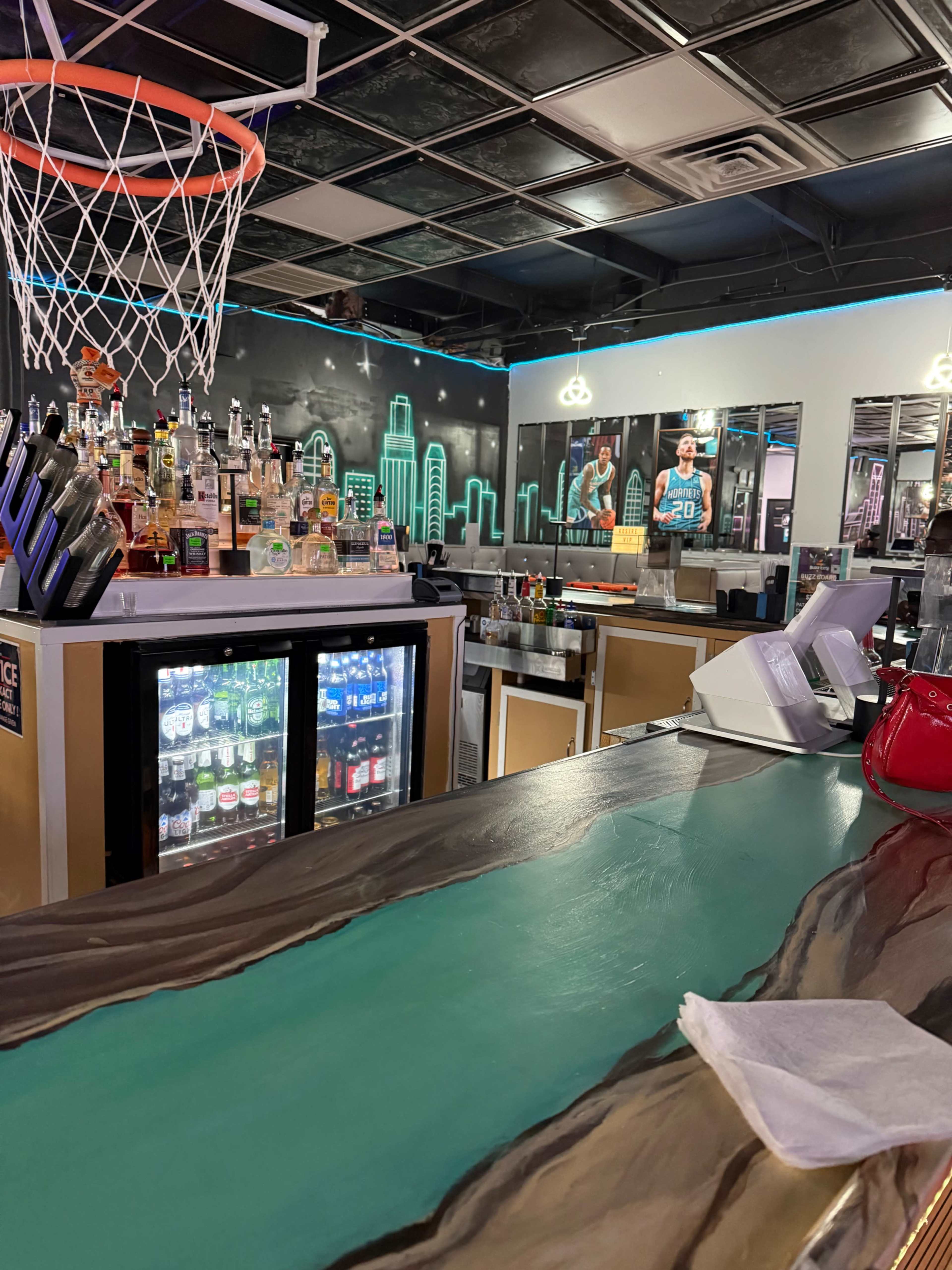 The image shows a bar area with a basketball hoop in the background, shelves of various bottles, and a cooler displaying drinks.