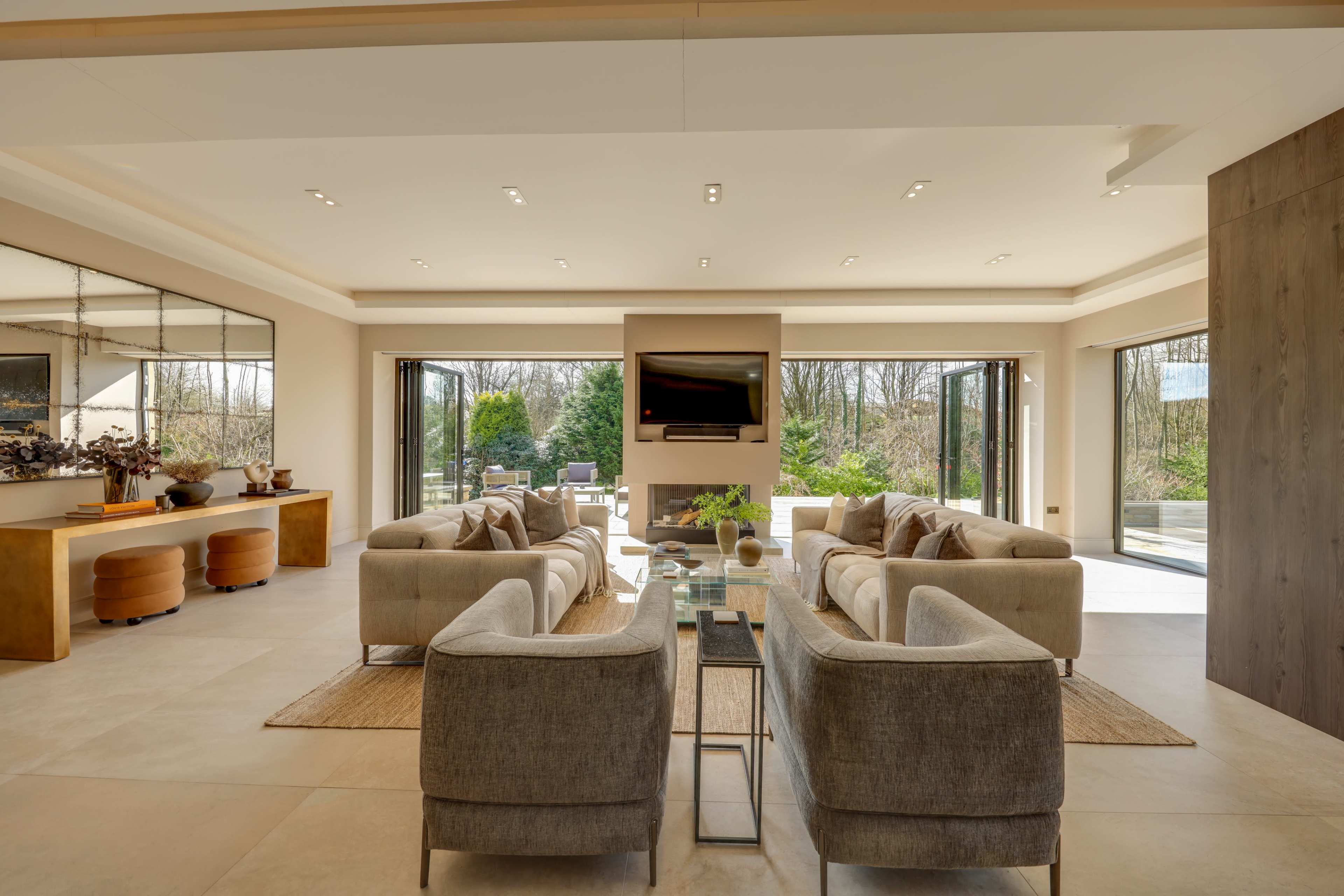 The image shows a modern living room with beige sofas arranged around a glass coffee table, large windows overlooking greenery, and a flat-screen TV mounted on a central wall.