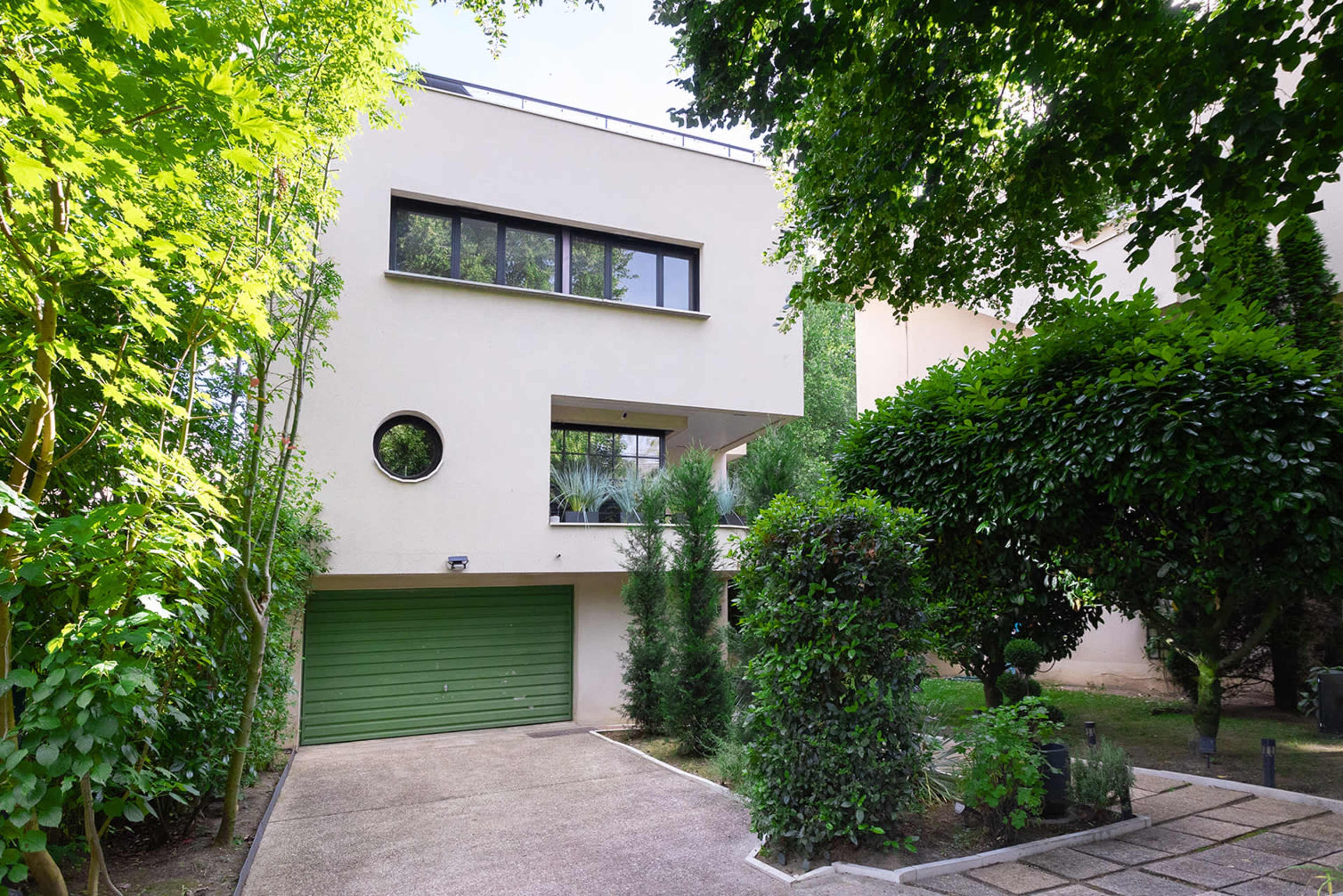 A modern two-story house with a circular window, a green garage door, and surrounded by trees and shrubs.