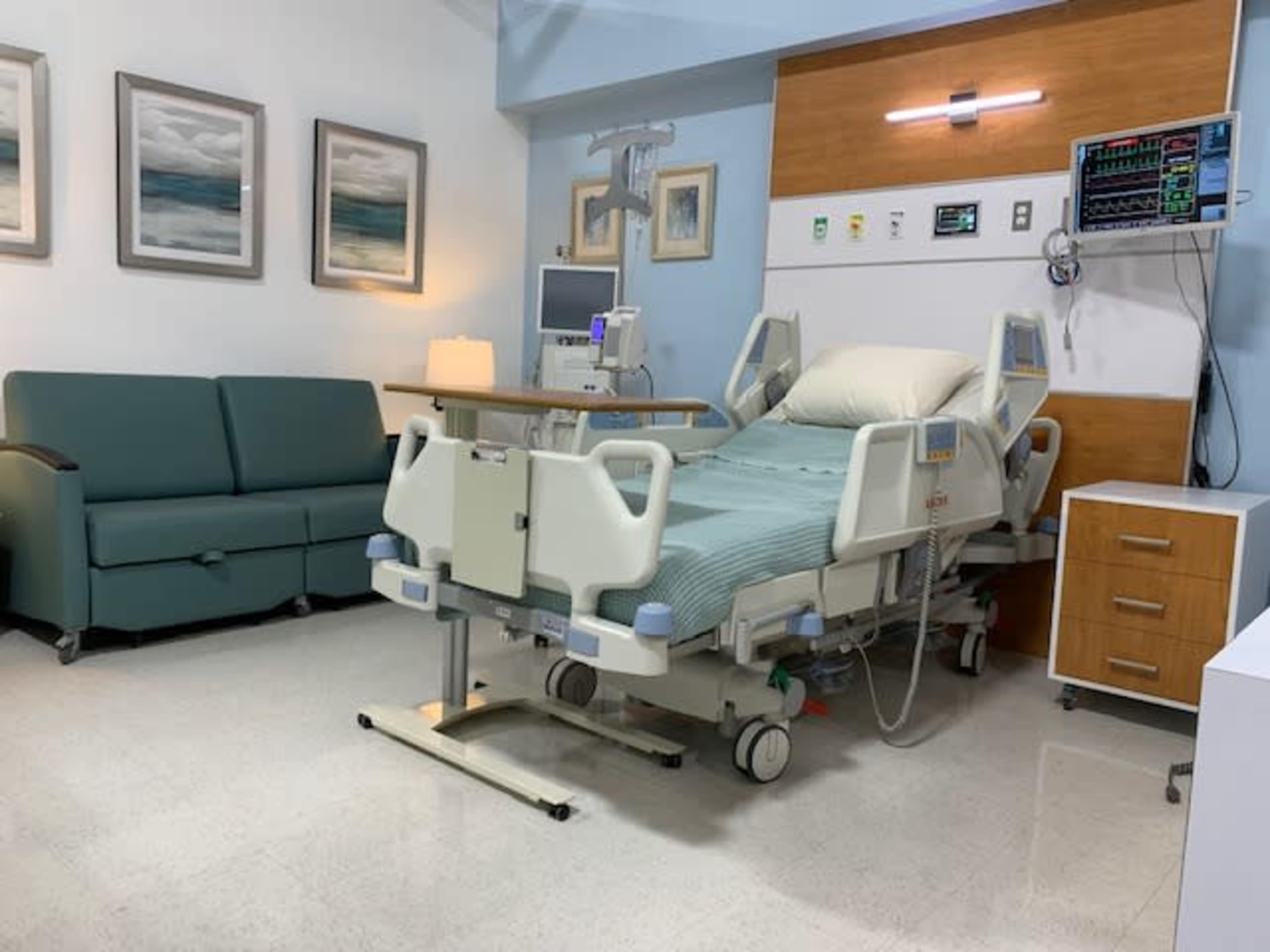 A hospital room with a bed, a couch, a nightstand, and medical equipment on the wall.