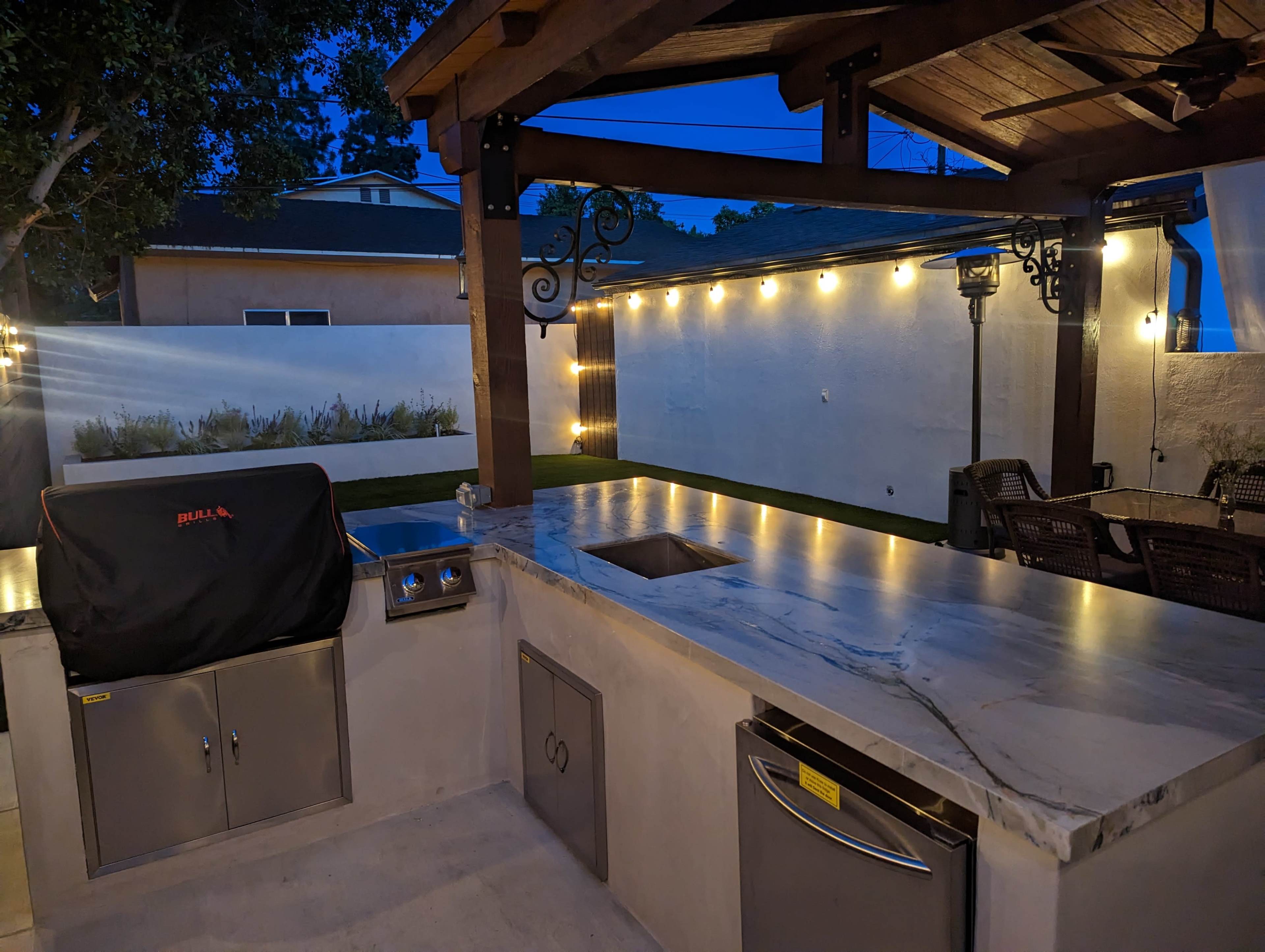 An outdoor kitchen area features a grill, sink, and countertop under a wooden pergola, with string lights illuminating the space at dusk.
