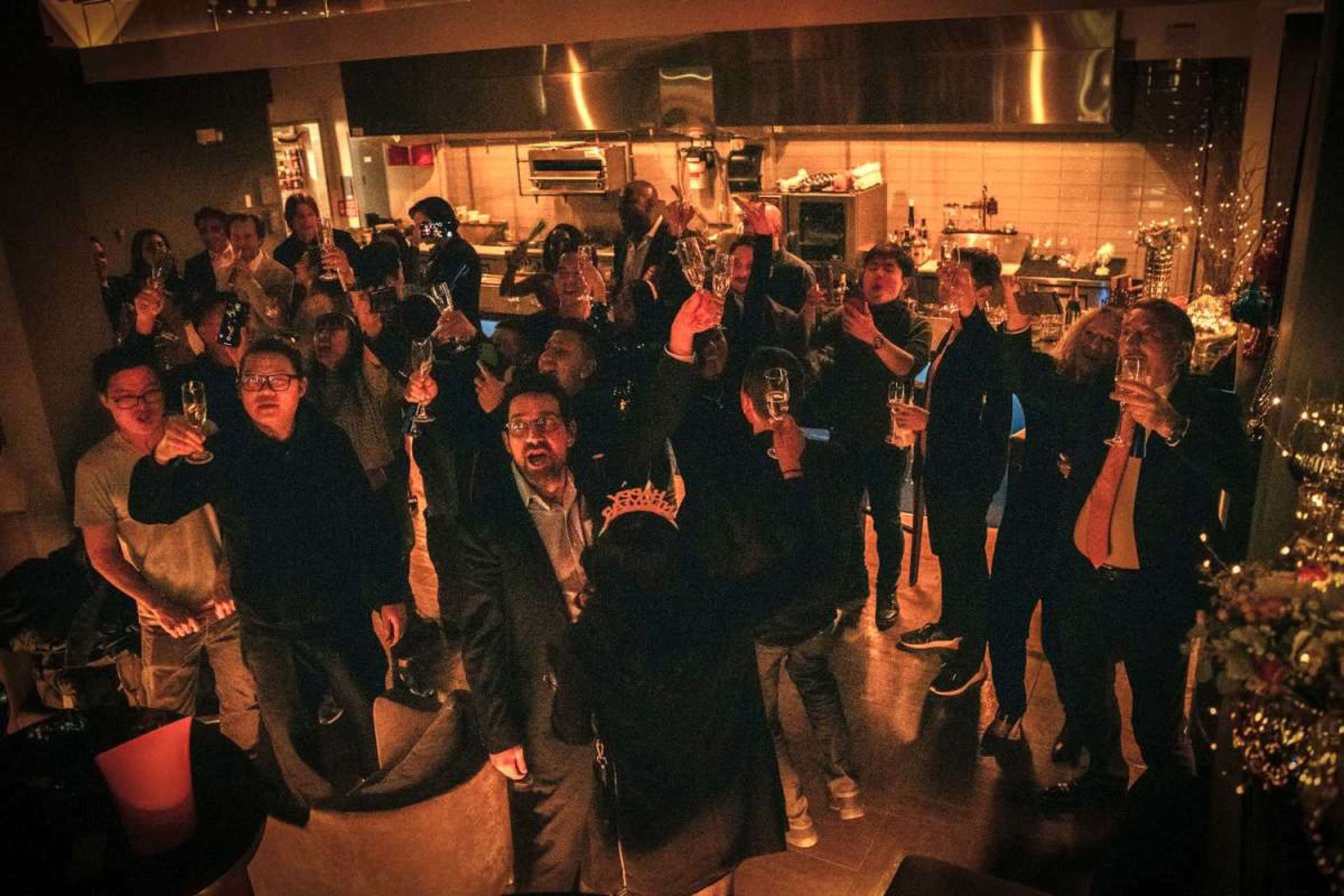 A group of people in formal attire raise glasses in a celebratory toast during a gathering in a dimly lit venue.