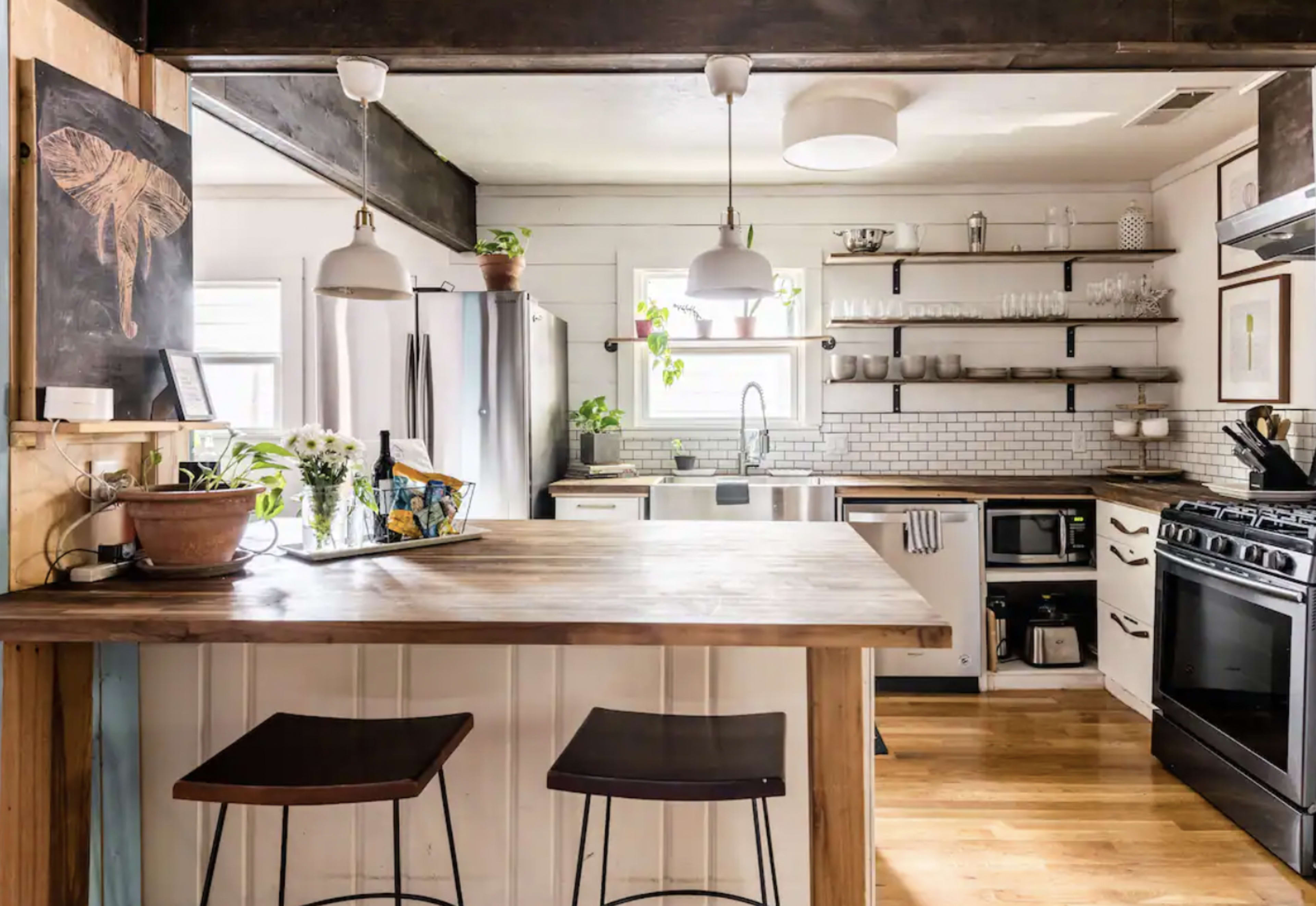 A modern kitchen featuring a wooden island with two stools, stainless steel appliances, open shelving, and a white tile backsplash.