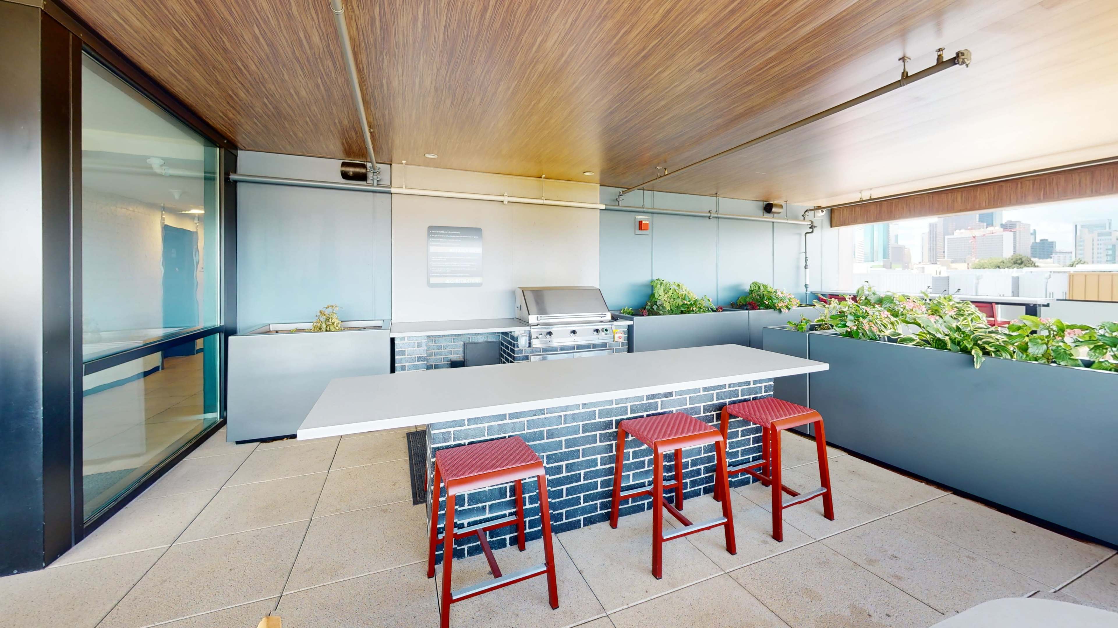The image shows a rooftop outdoor kitchen area featuring a grill, a long countertop, and three red stools against a backdrop of city buildings.