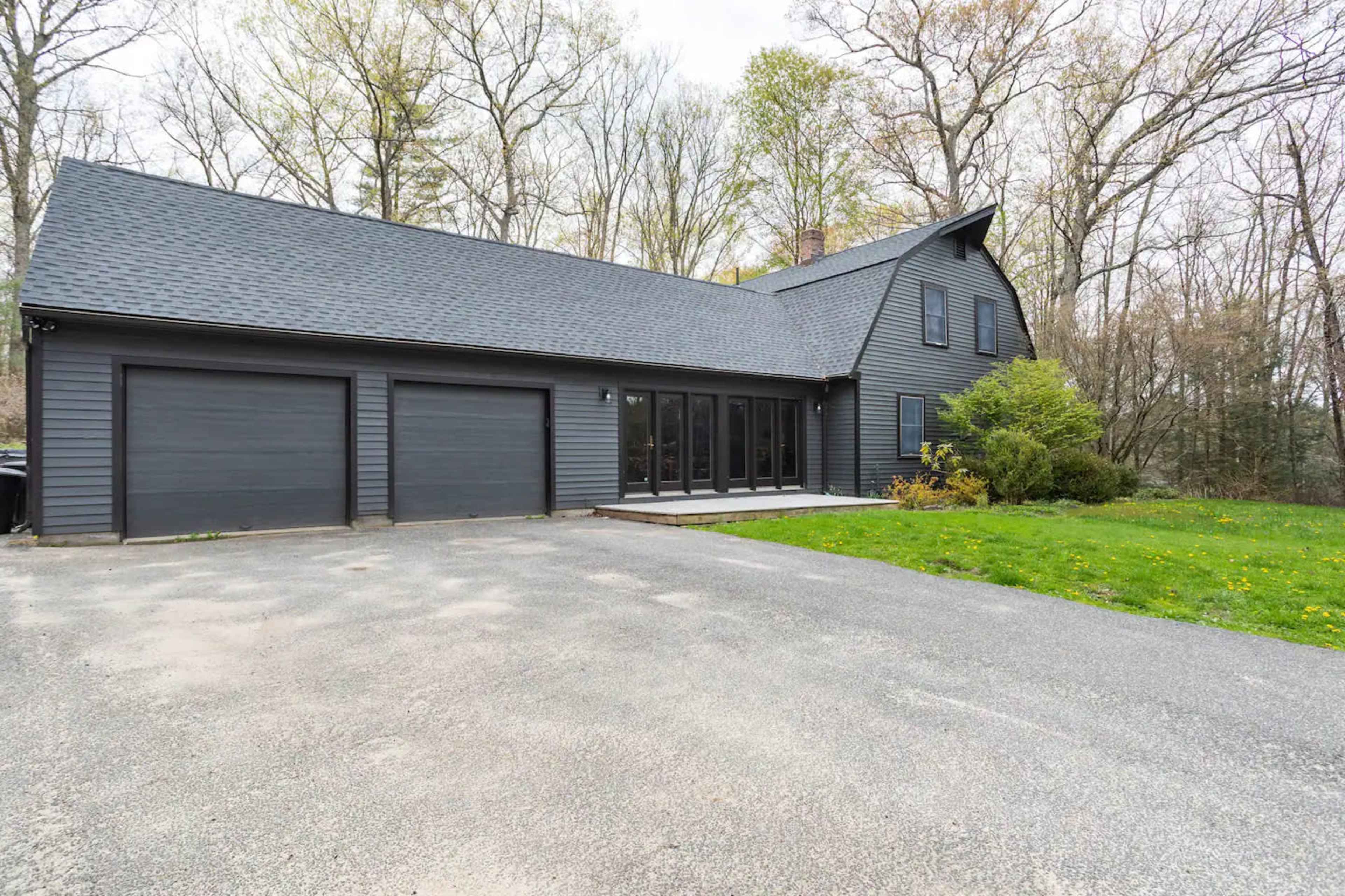 A black, two-story house with a steep roof and two attached garages is set against a backdrop of trees and grass.