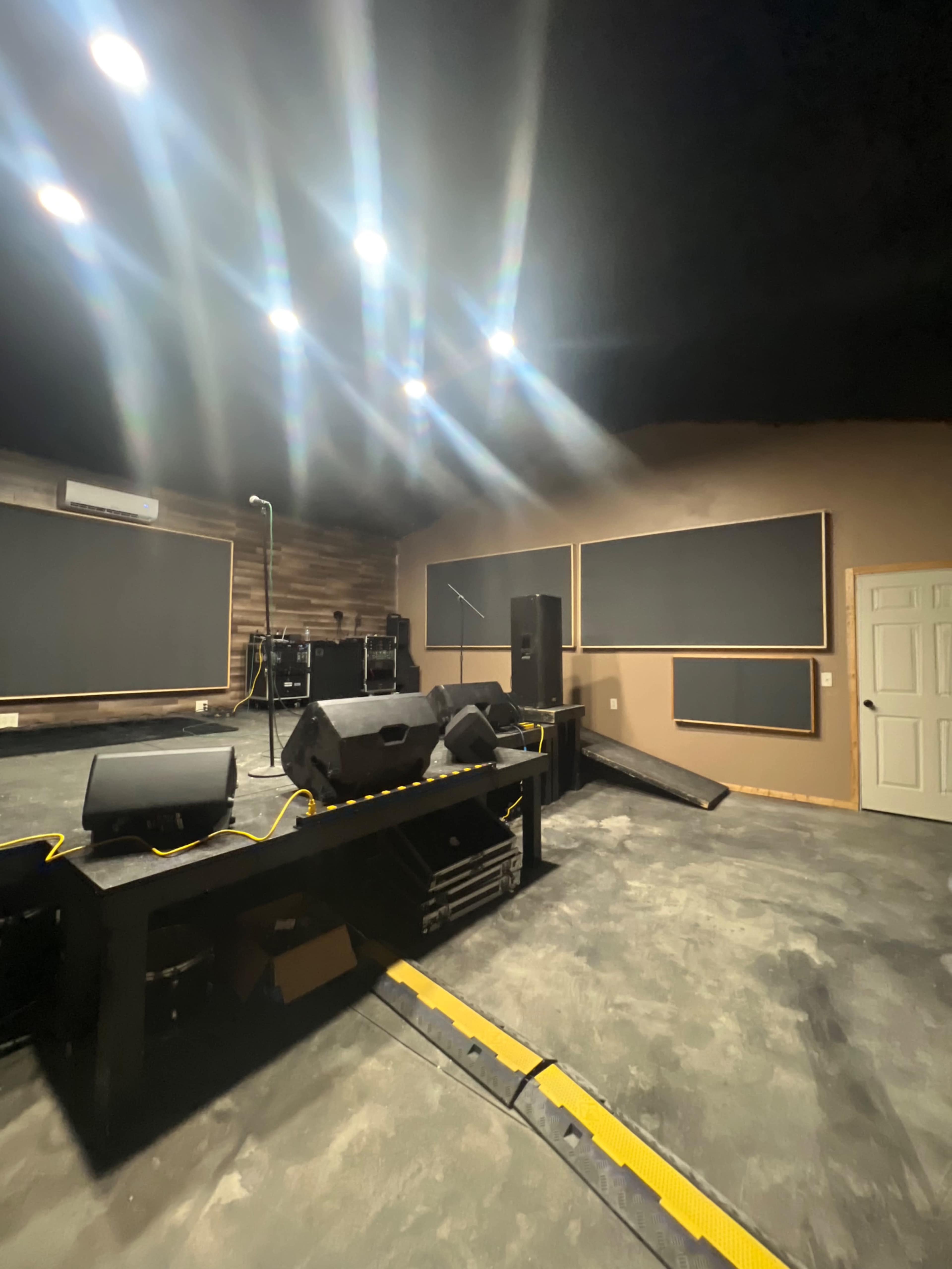 A dimly lit performance space featuring a stage with speakers and a microphone setup, surrounded by large blank sound-absorption panels on the walls.