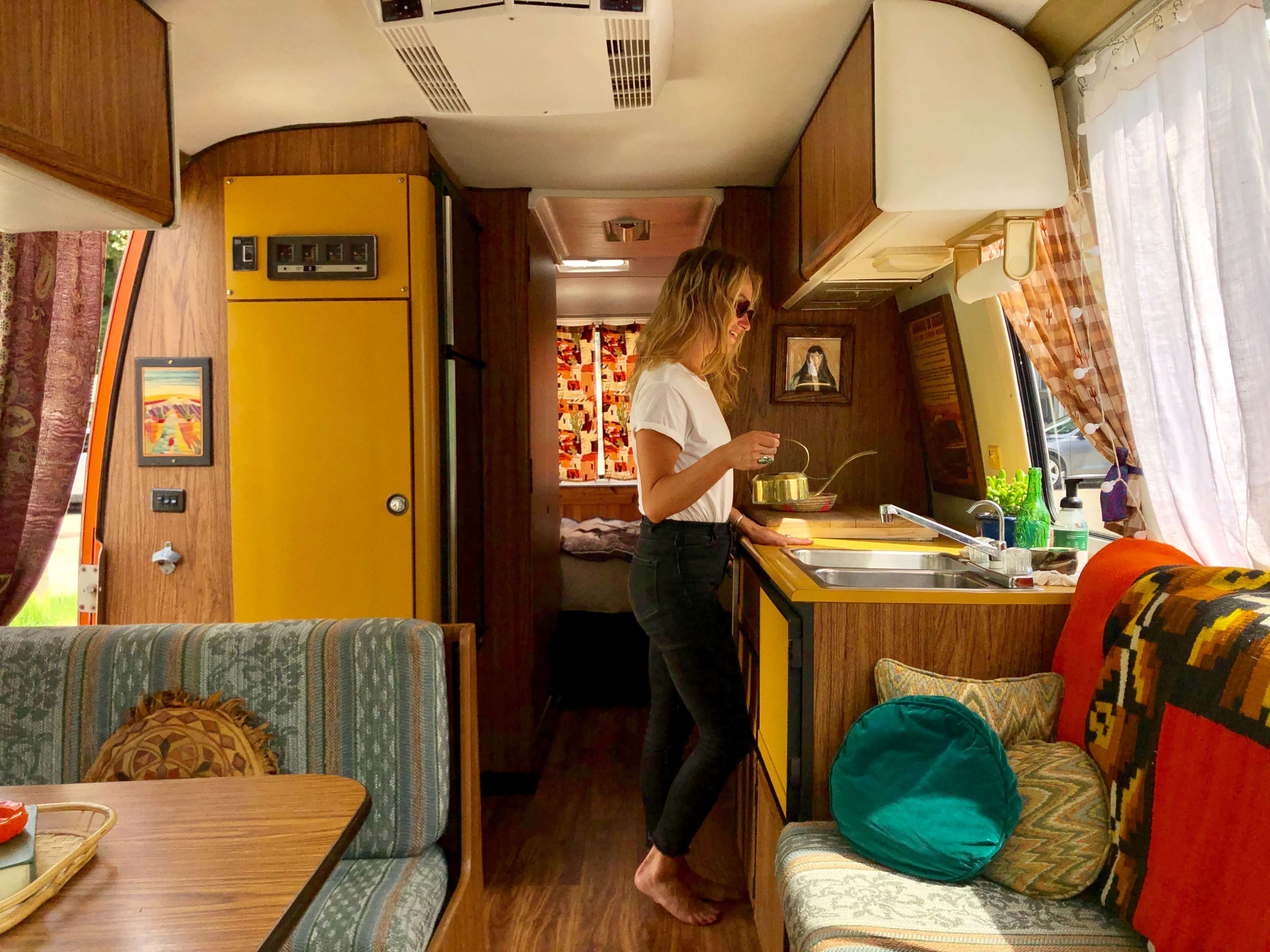 A woman stands in a retro-styled camper van, preparing food at a kitchen counter with vibrant decor.