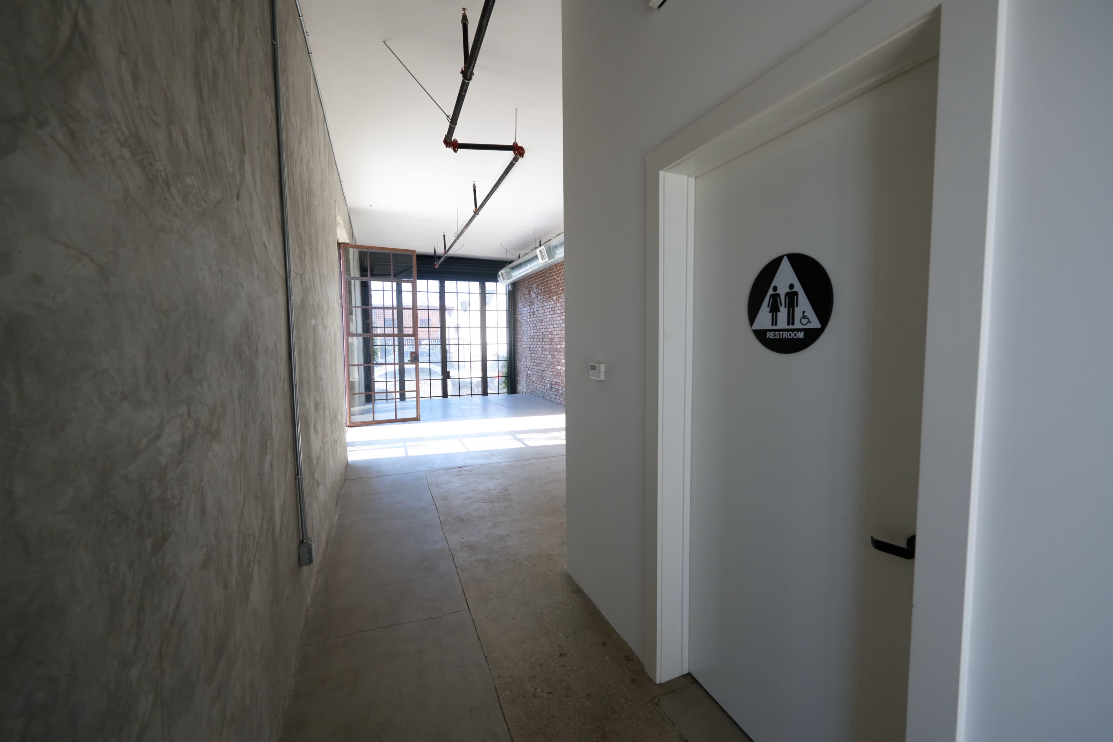 The image shows a hallway leading to a restroom door, with large windows and a concrete wall in the background.