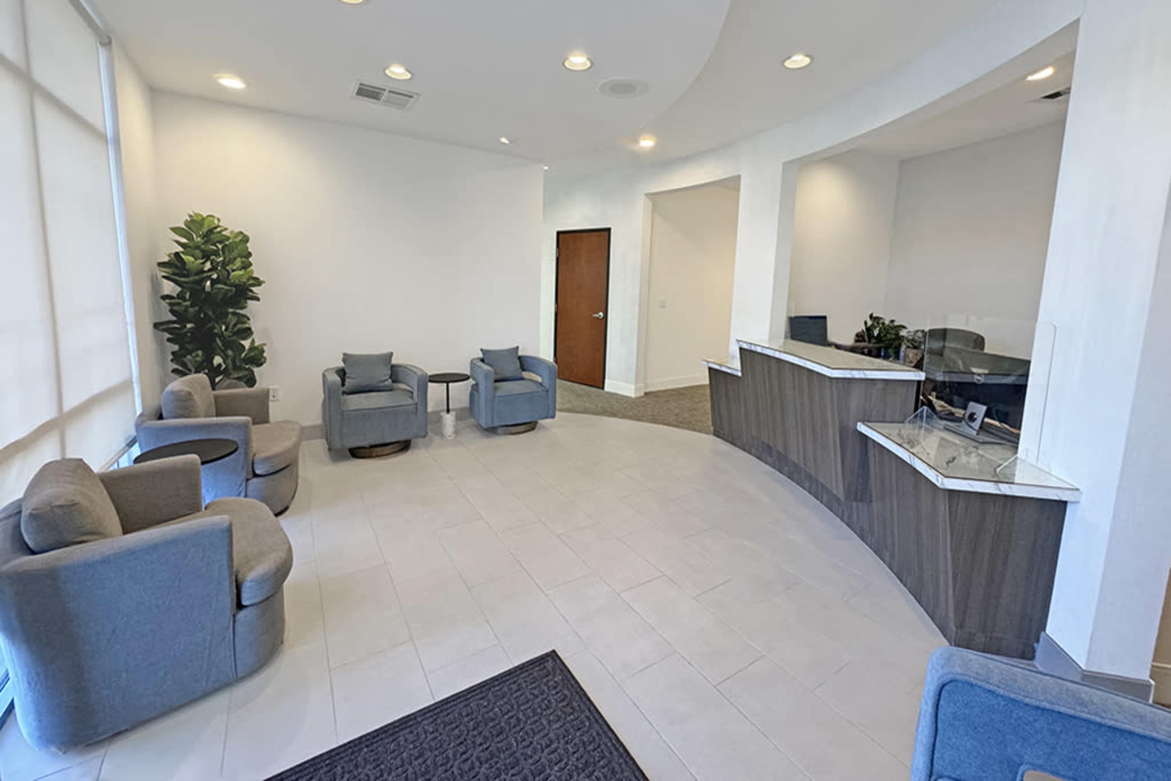 The image shows a modern waiting area with gray upholstered chairs and a reception desk against a light-colored wall.