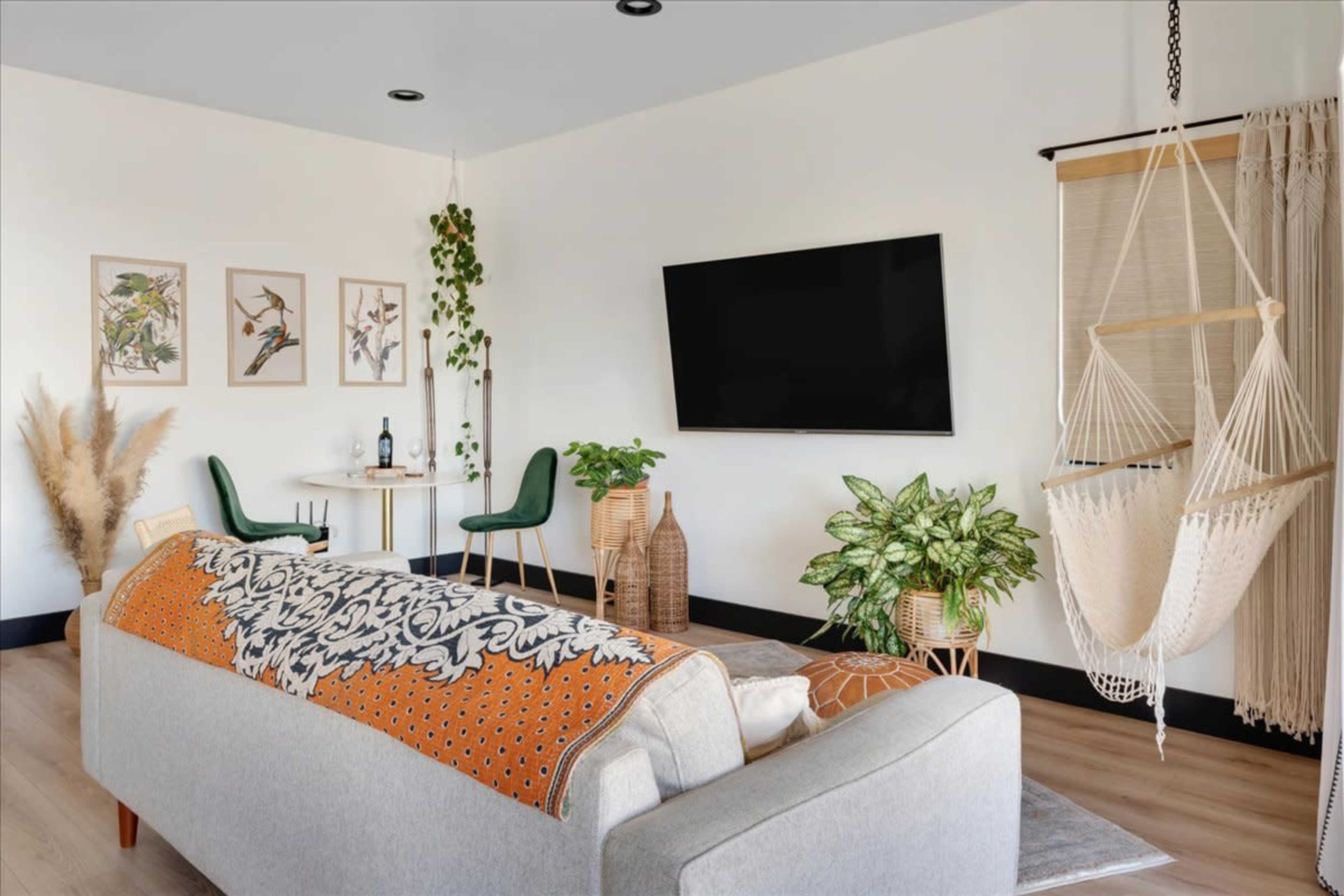 The image shows a modern living room featuring a light gray sofa, a wall-mounted television, a small dining area with green chairs, and a hammock chair beside decorative plants.