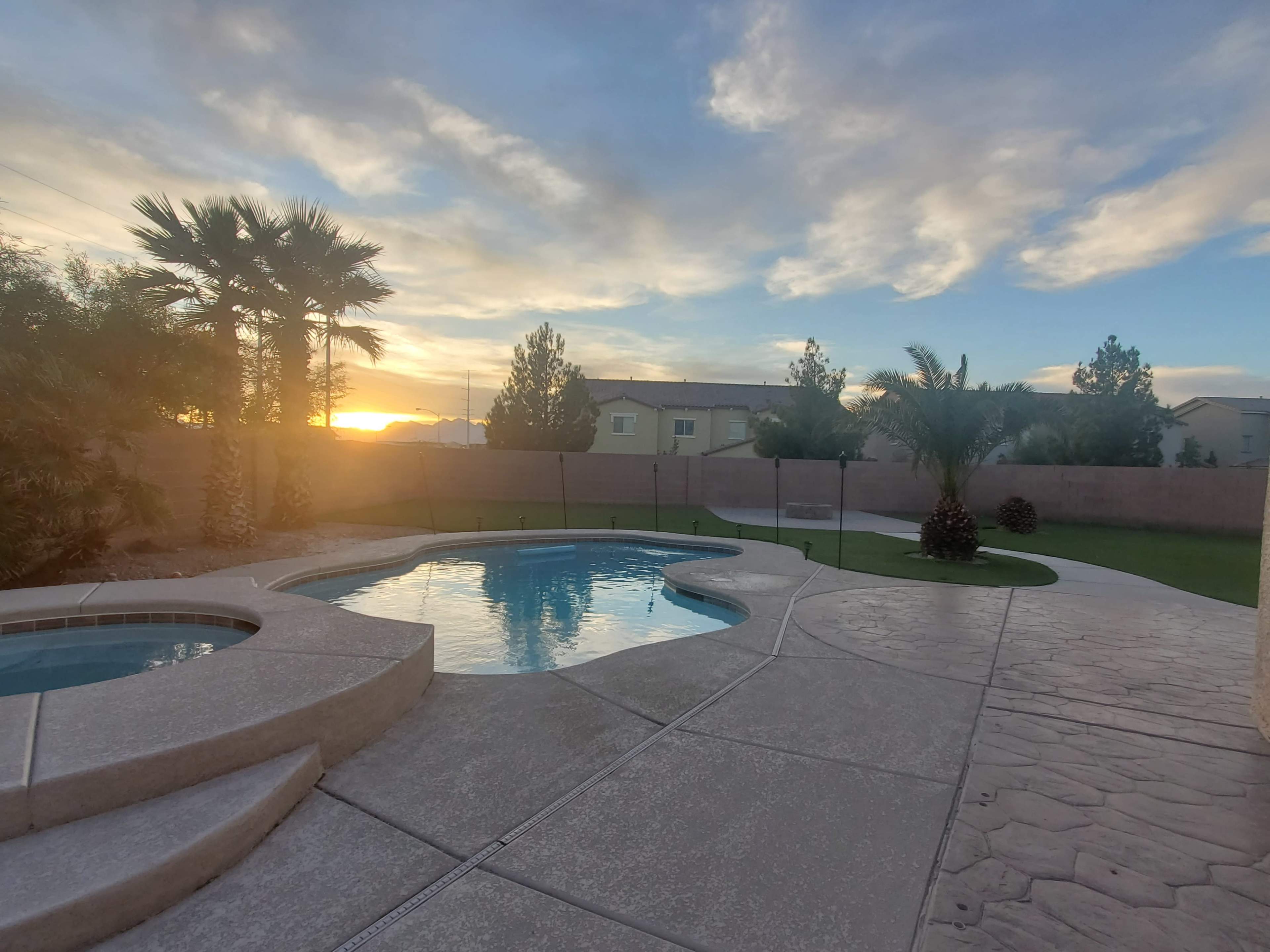 The image shows a backyard with a swimming pool surrounded by palm trees and a grassy area during sunset.