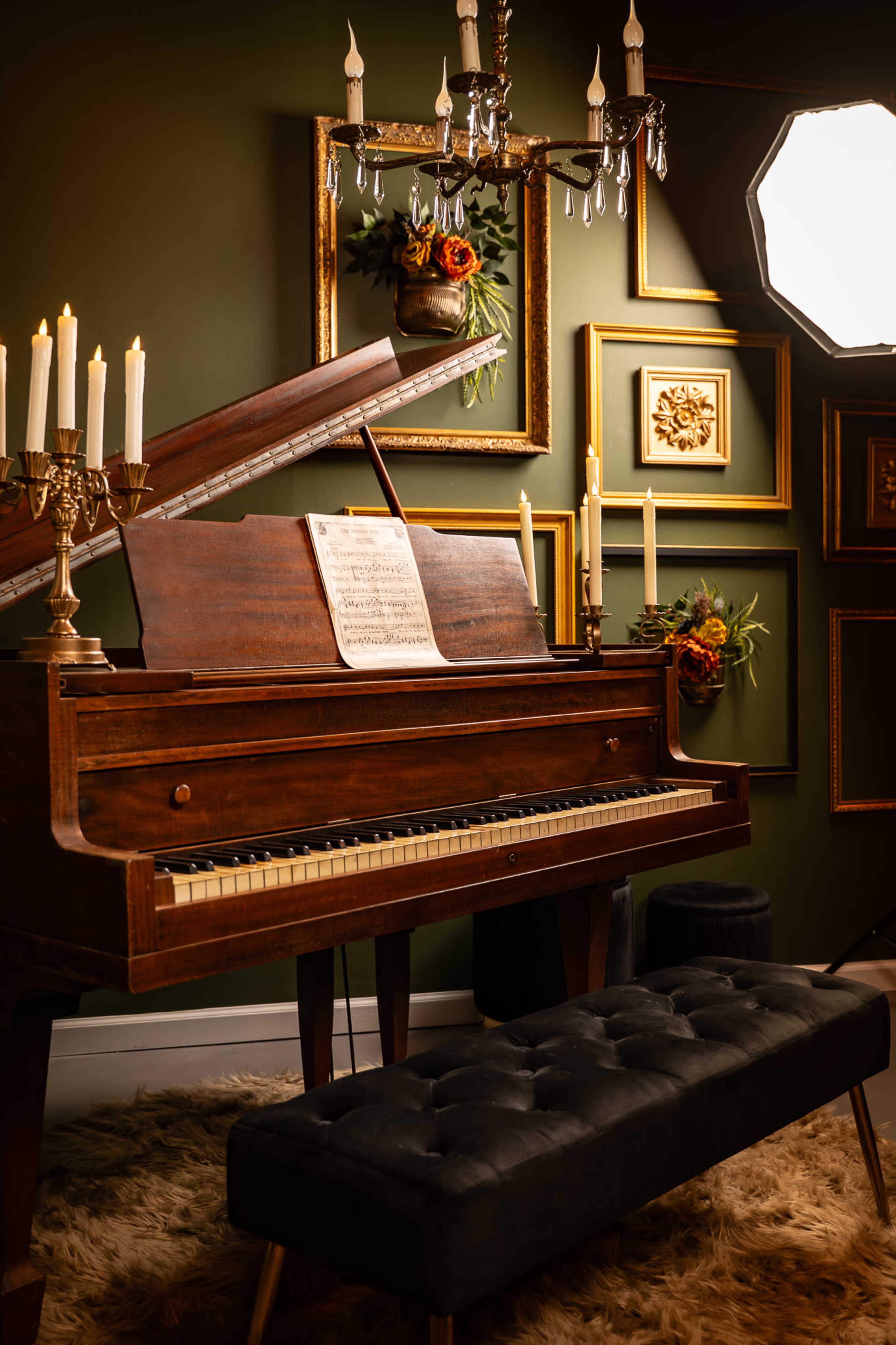 A vintage wooden piano is positioned beside a chandelier and adorned with decorative candles, surrounded by framed artwork on a green wall.