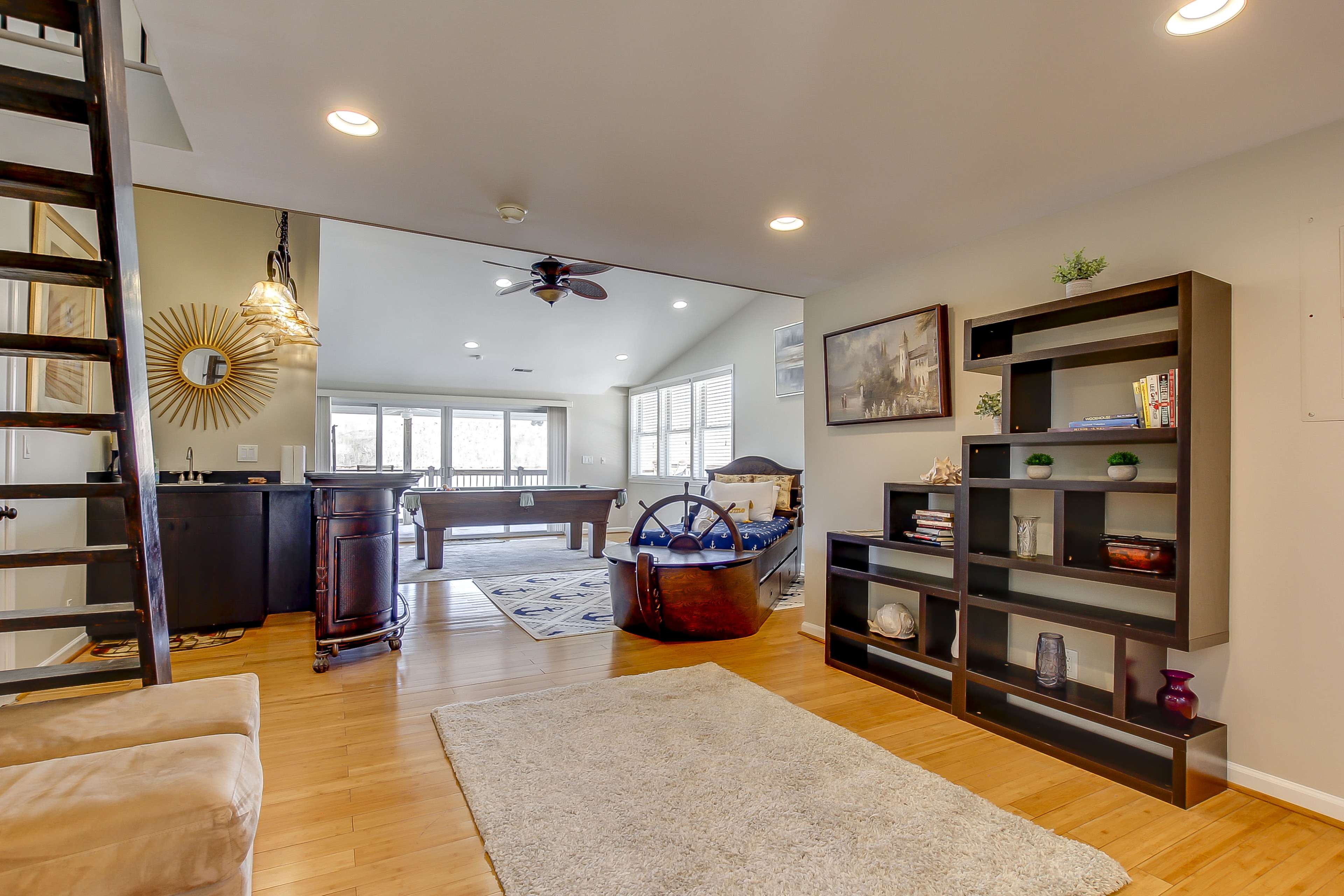 A modern living space featuring a wooden staircase, a black bookshelf, and large windows overlooking a pool table and a balcony area.