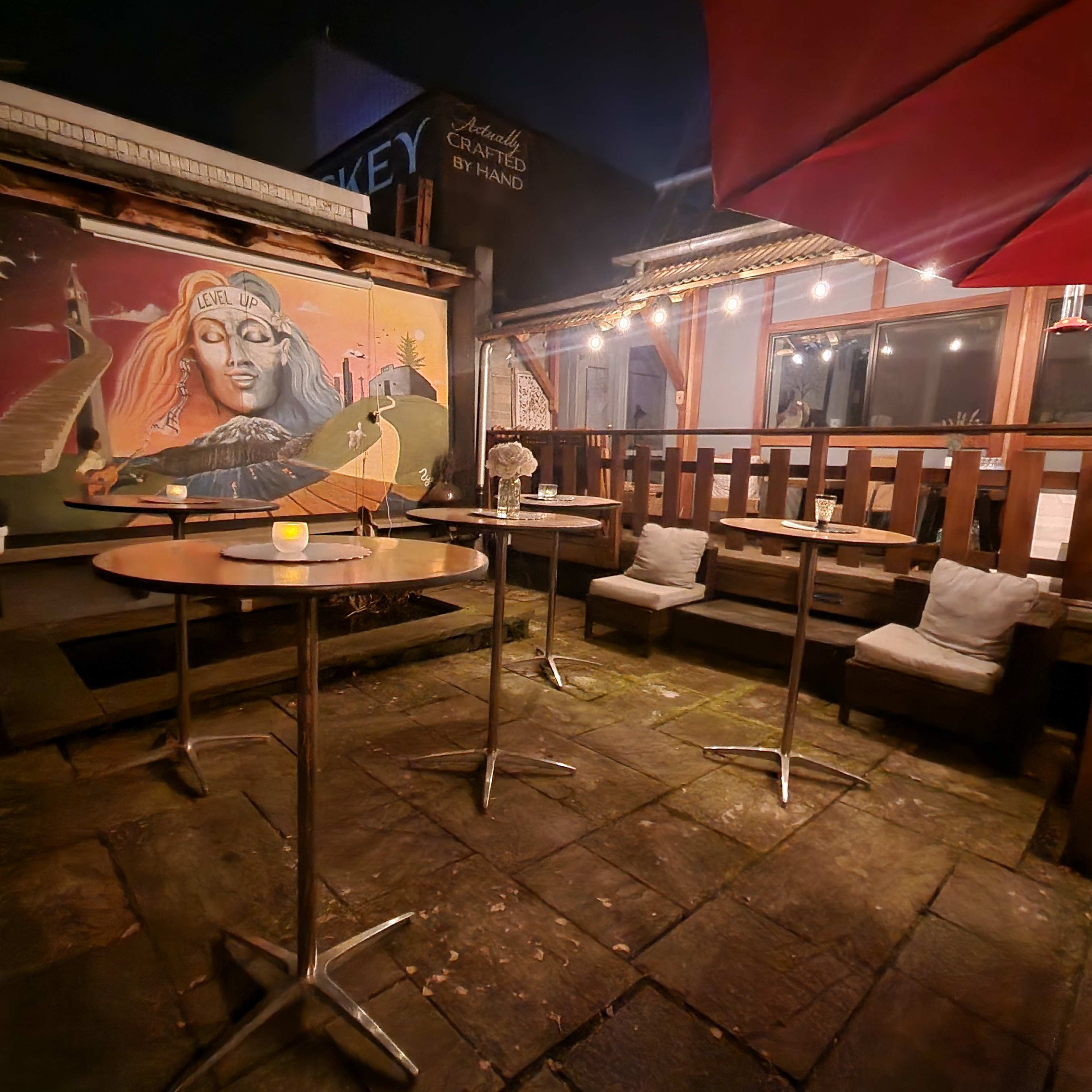 The scene shows an outdoor bar area at night with several empty high tables, string lights overhead, and a mural featuring a woman on the back wall.