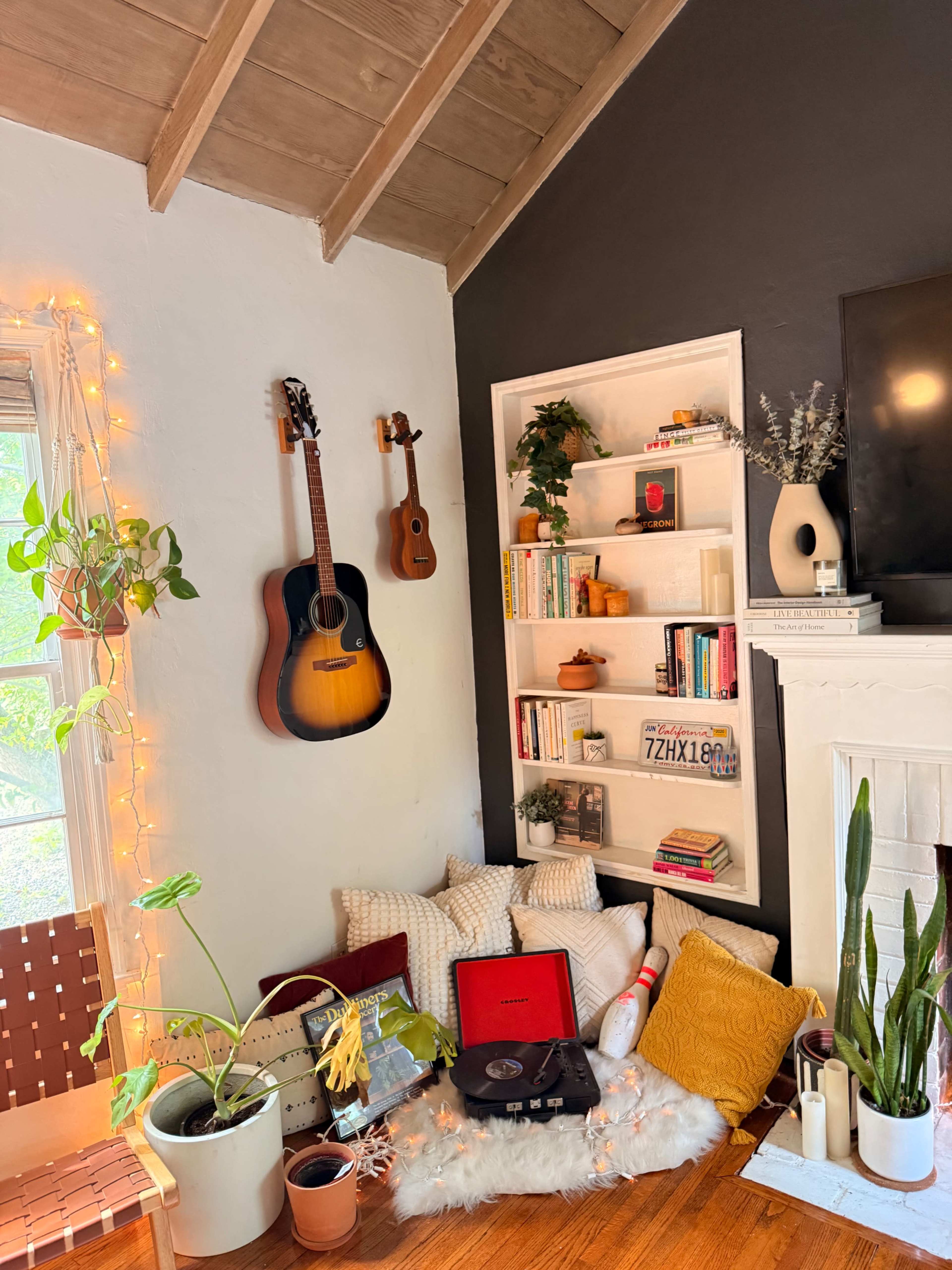 A cozy corner of a living room features an acoustic guitar and ukulele on the wall, a bookshelf filled with books and decorative items, a record player on the floor among cushions, and potted plants.