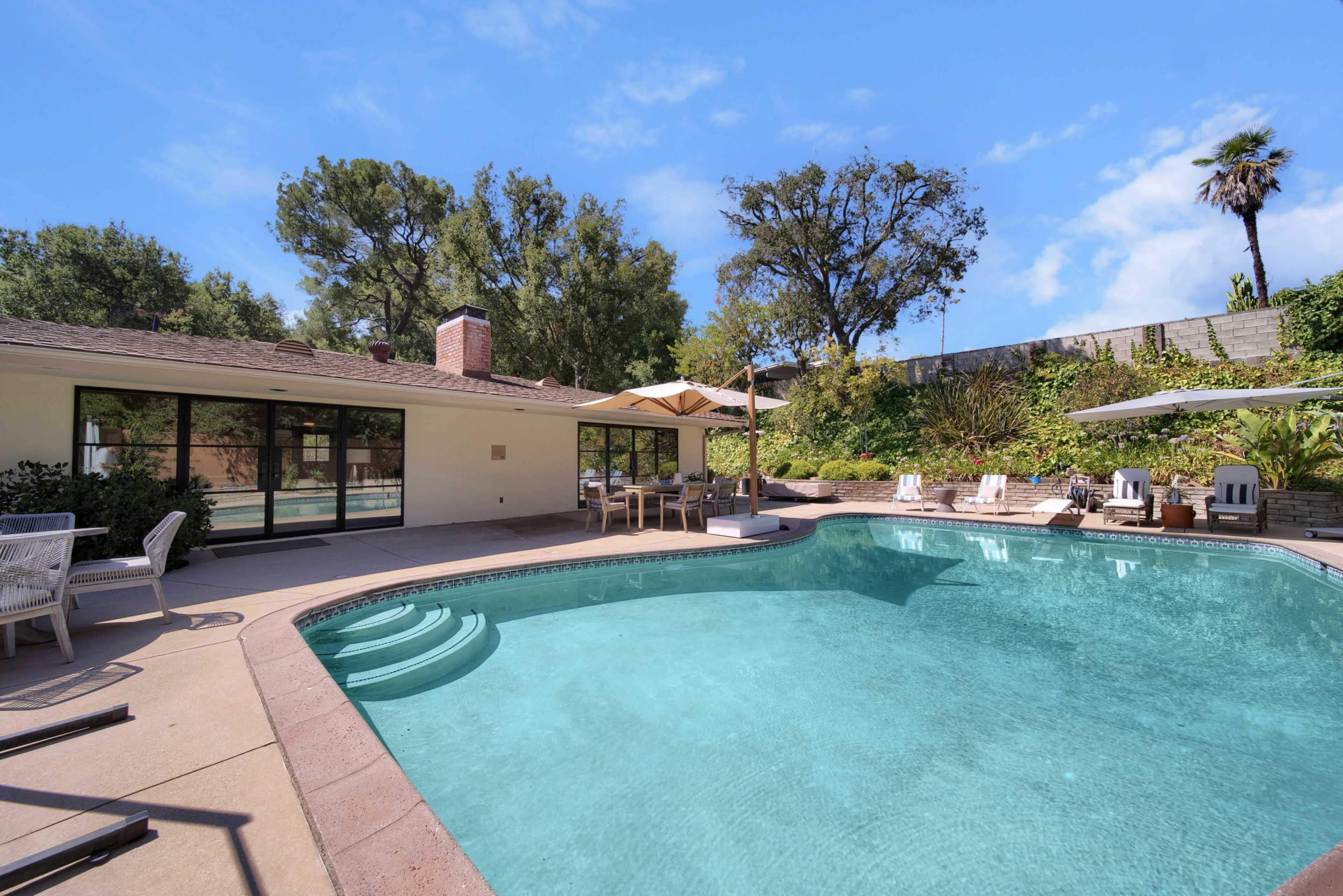 The image shows a swimming pool surrounded by lounge chairs and a dining area, with lush greenery and trees in the background.