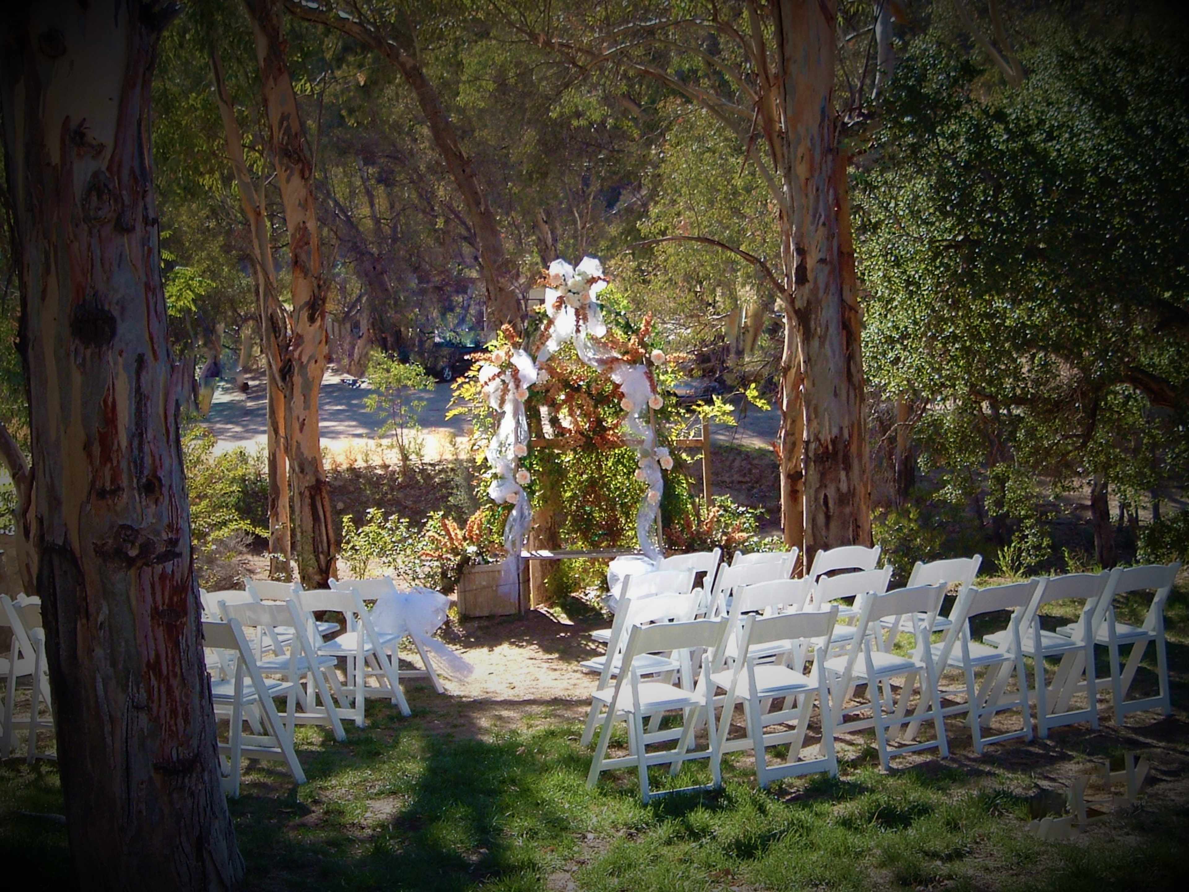 A decorated outdoor wedding altar is set up among trees, facing rows of white chairs arranged for guests.