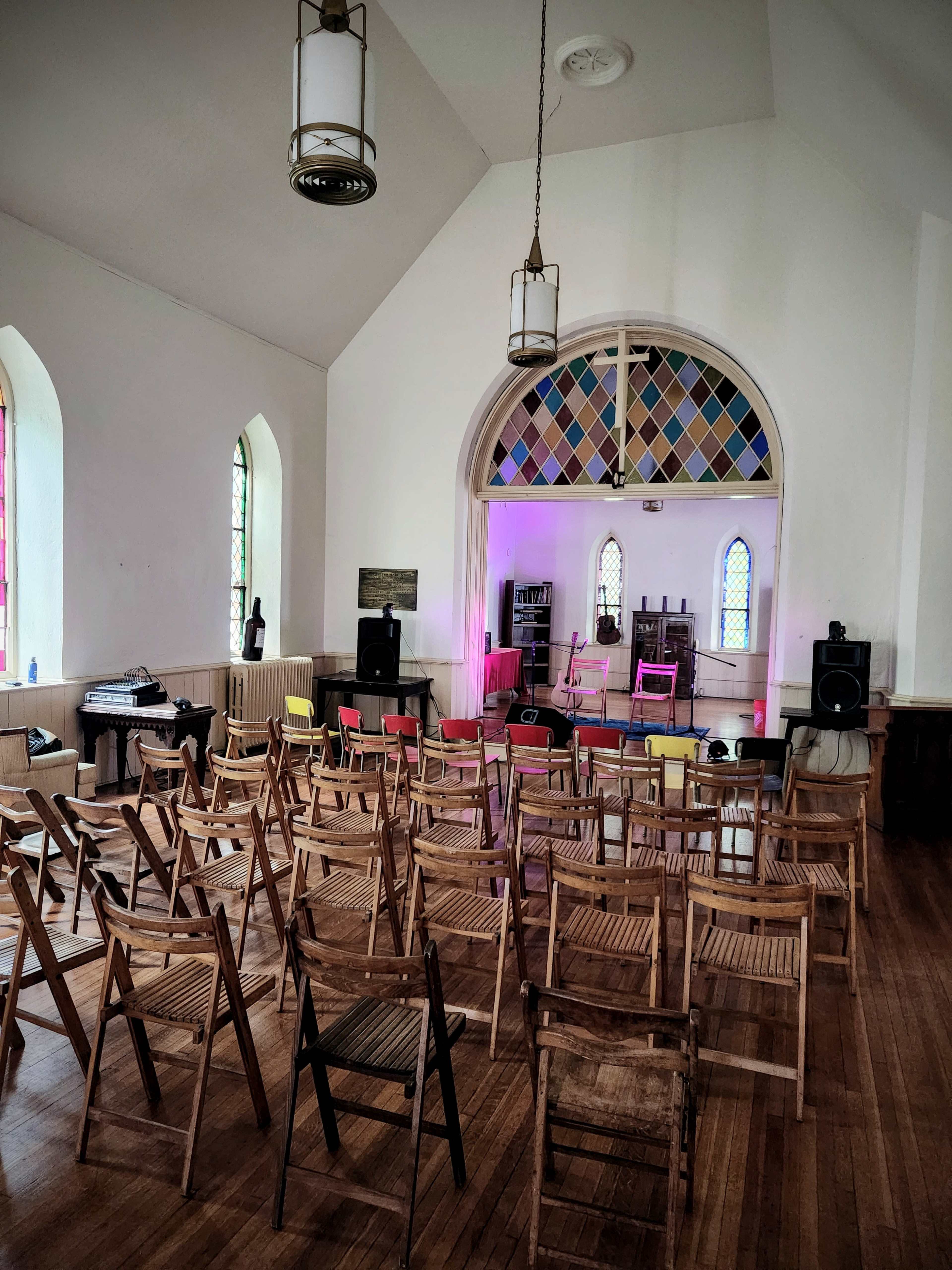 A sparsely decorated room with folding chairs arranged in rows, a small stage at the front, and colorful lights illuminating the space.