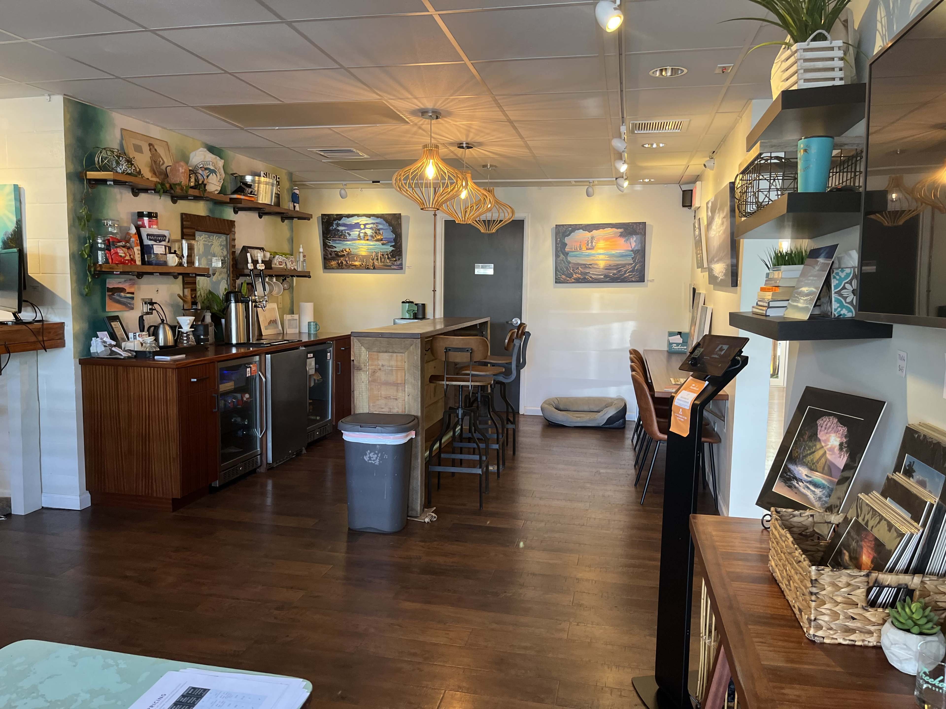 An inviting café interior with a wooden counter, seating area, shelves displaying art, and a small refrigerator.