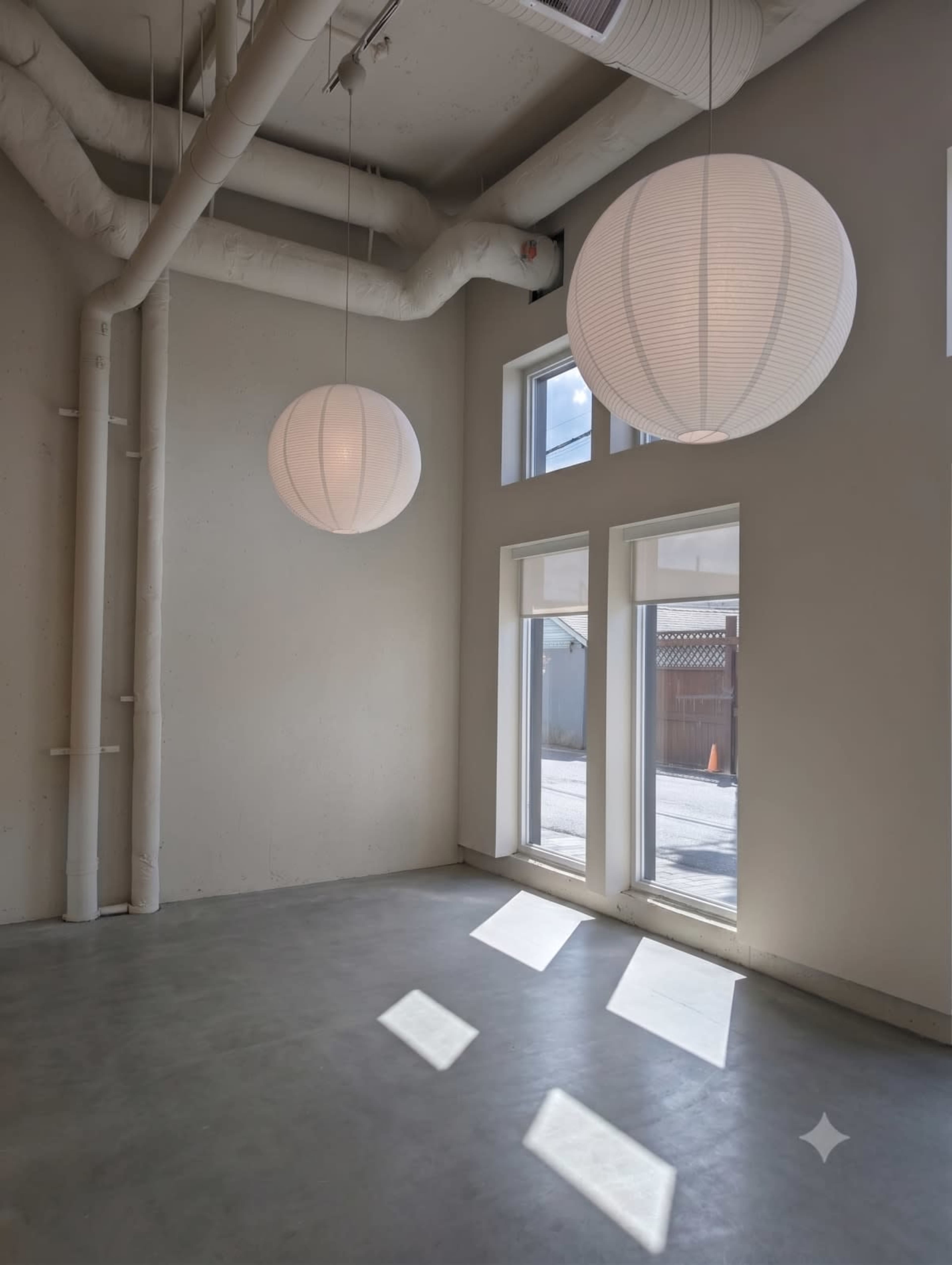 The image shows a spacious room with large windows, two round paper lanterns hanging from the ceiling, and light casting shadows on the concrete floor.