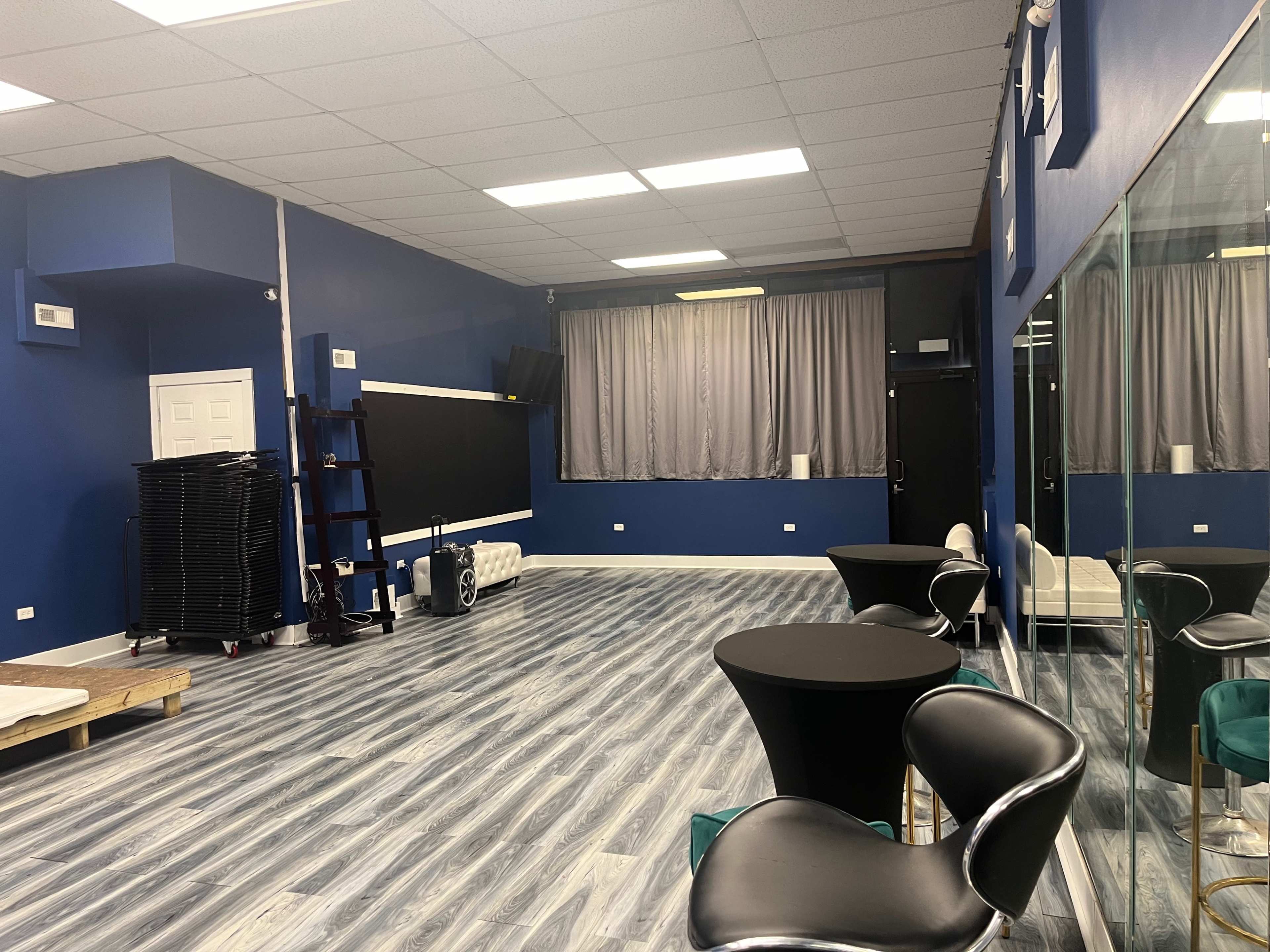 A spacious, empty room with blue walls, gray wooden flooring, a large mirror along one wall, and two black tables with chairs.