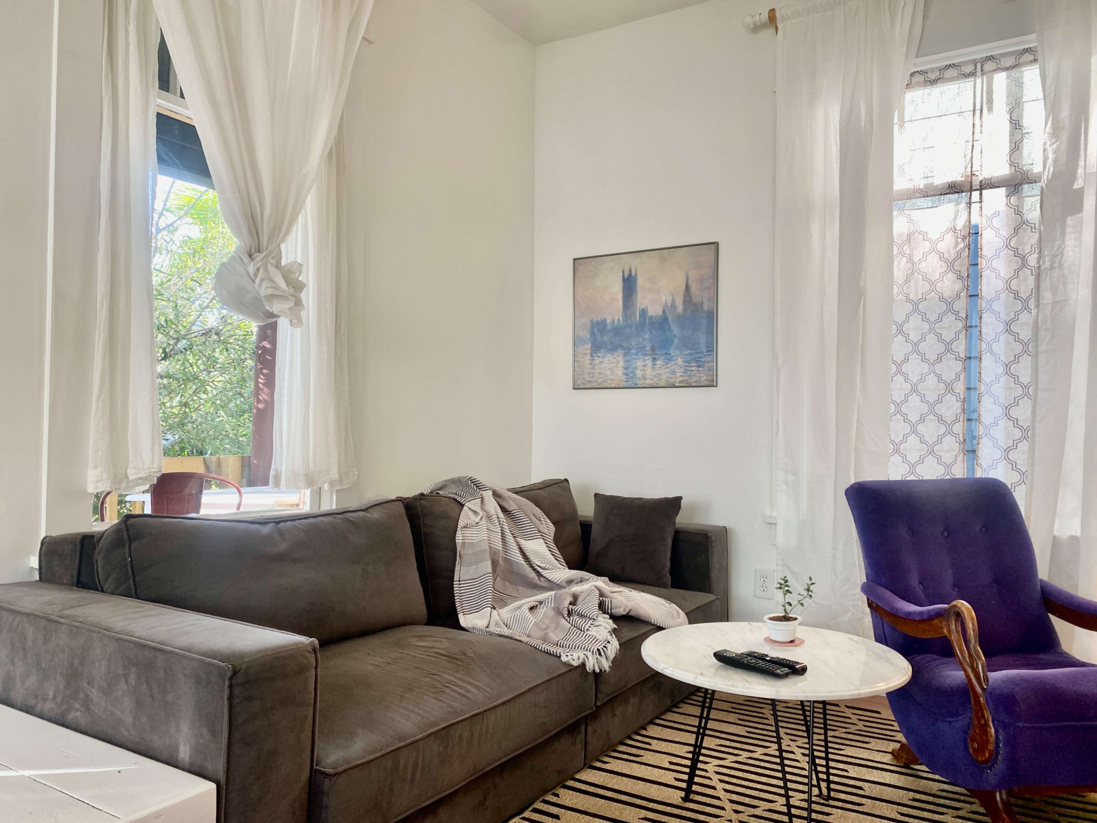 A living room with a dark gray sofa, a purple armchair, a round white coffee table, and large windows with sheer white curtains.
