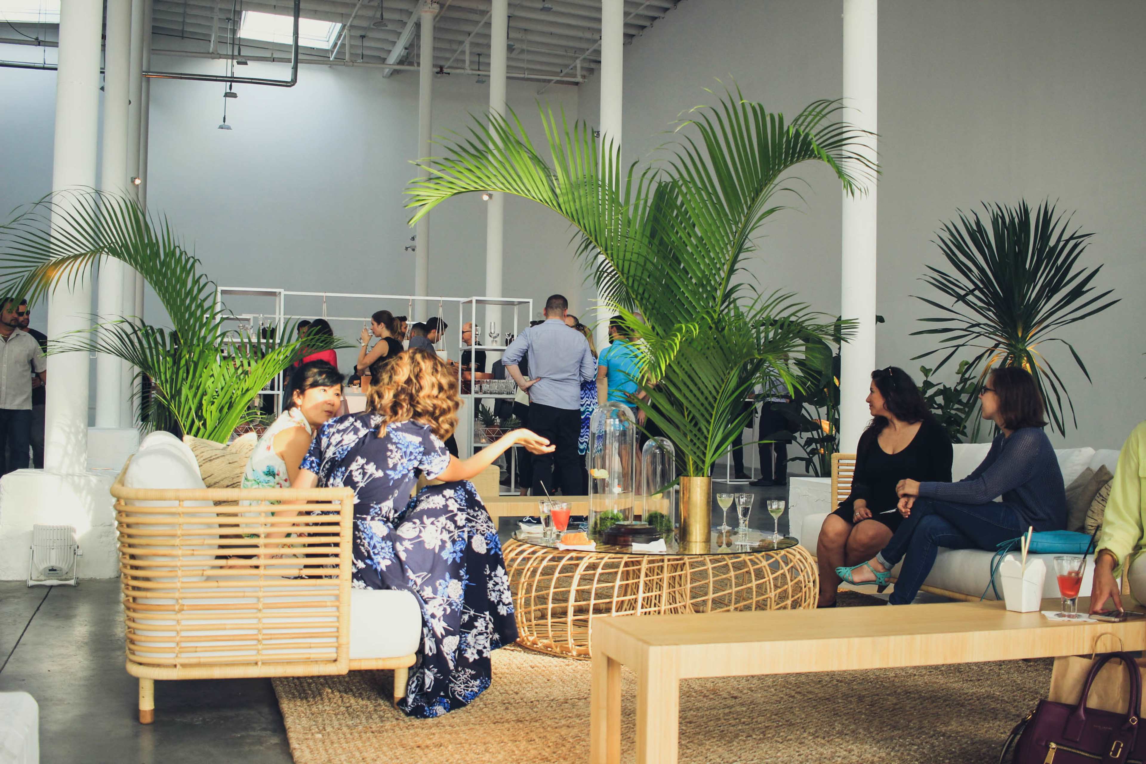A group of people socializes in a modern, open space furnished with a rattan seating area and tropical plants.