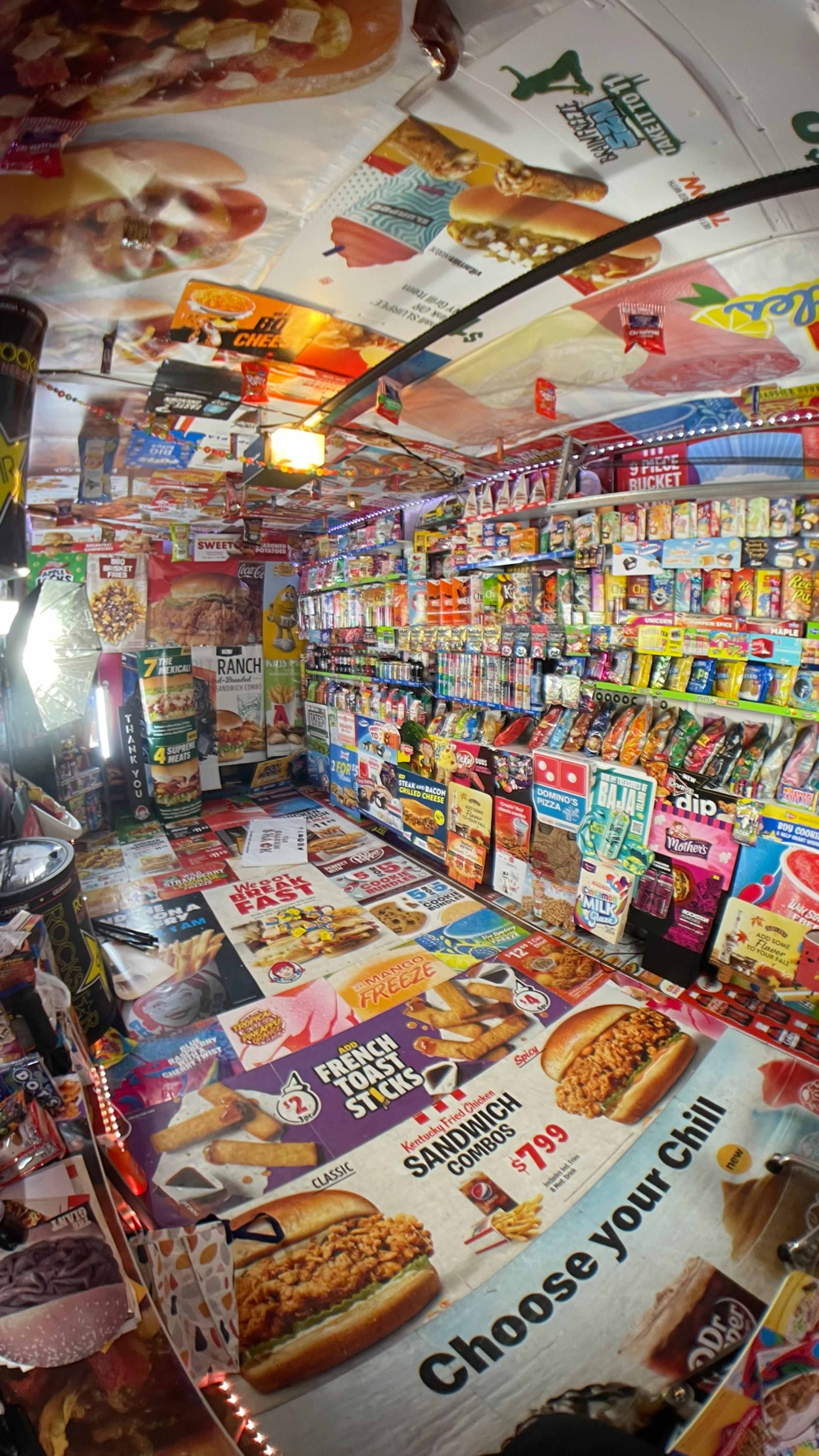 The image shows the interior of a convenience store filled with various snacks and food items displayed on the walls, shelves, and floor, all surrounded by colorful advertisements.