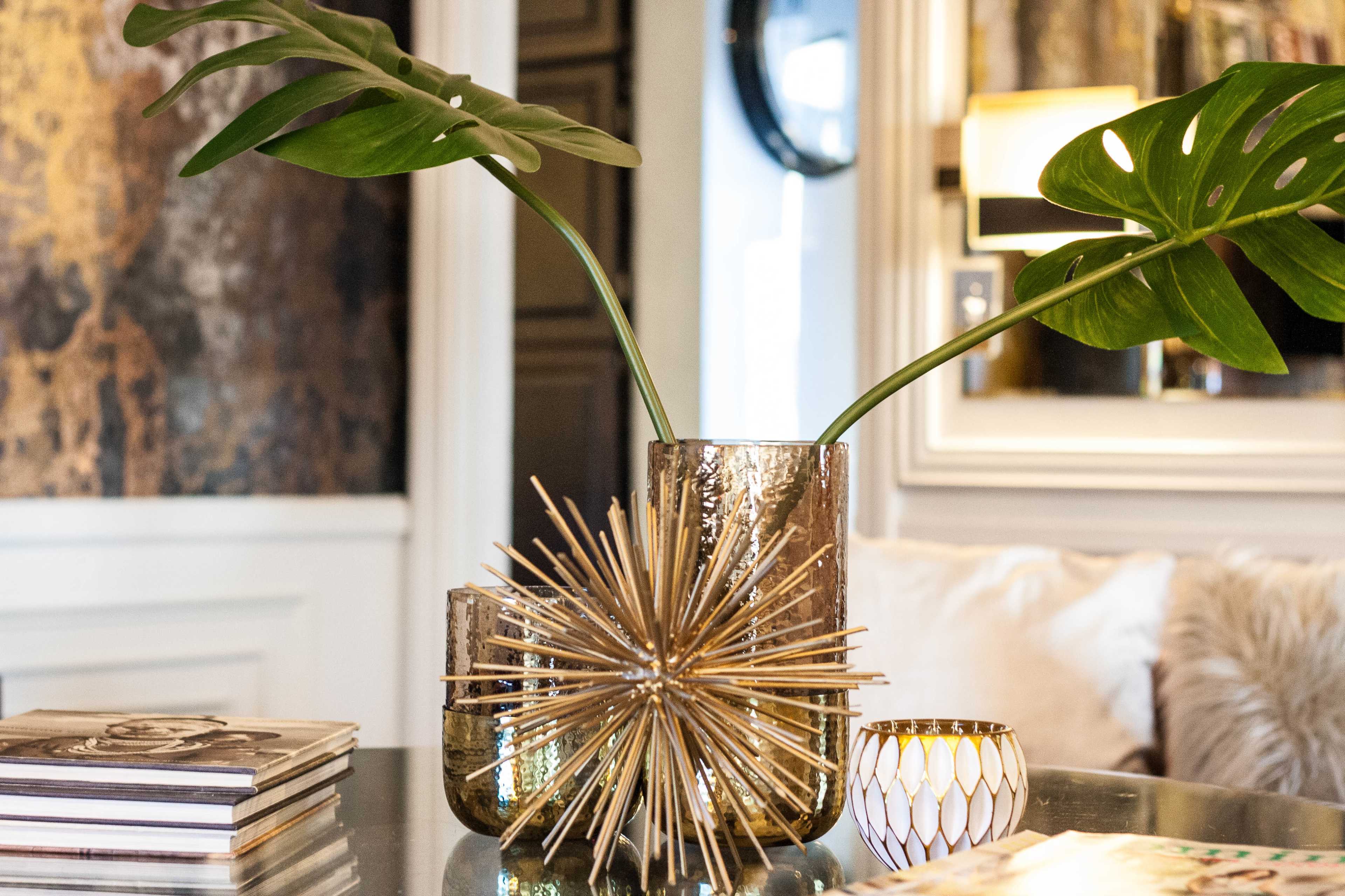 A decorative arrangement features two metallic vases with tropical leaves and a starburst sculpture on a coffee table surrounded by books and a candle.
