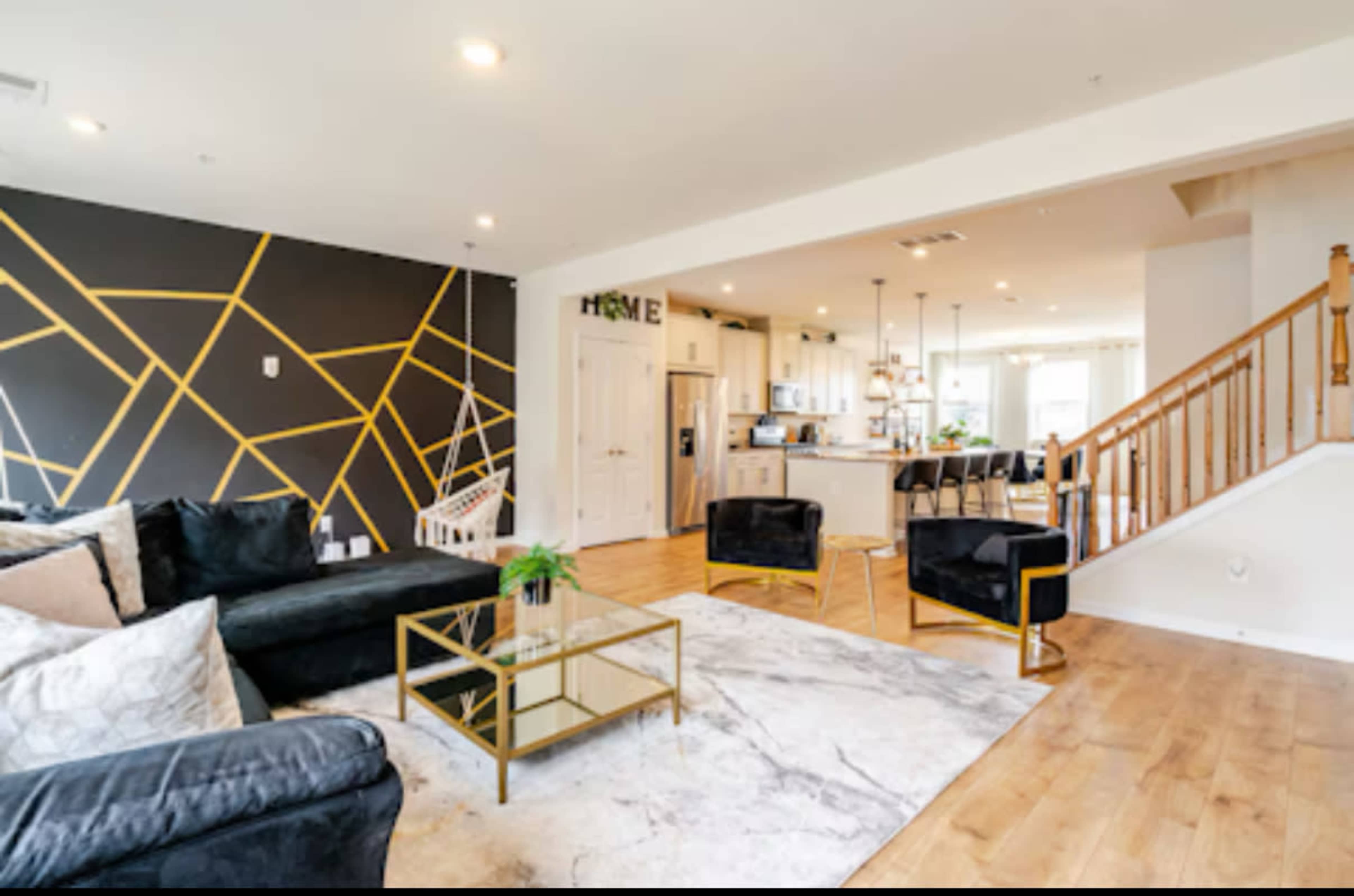 The image shows a modern living room with a geometric black and gold accent wall, a sectional sofa, and a marble rug, leading to a kitchen area with bar stools.