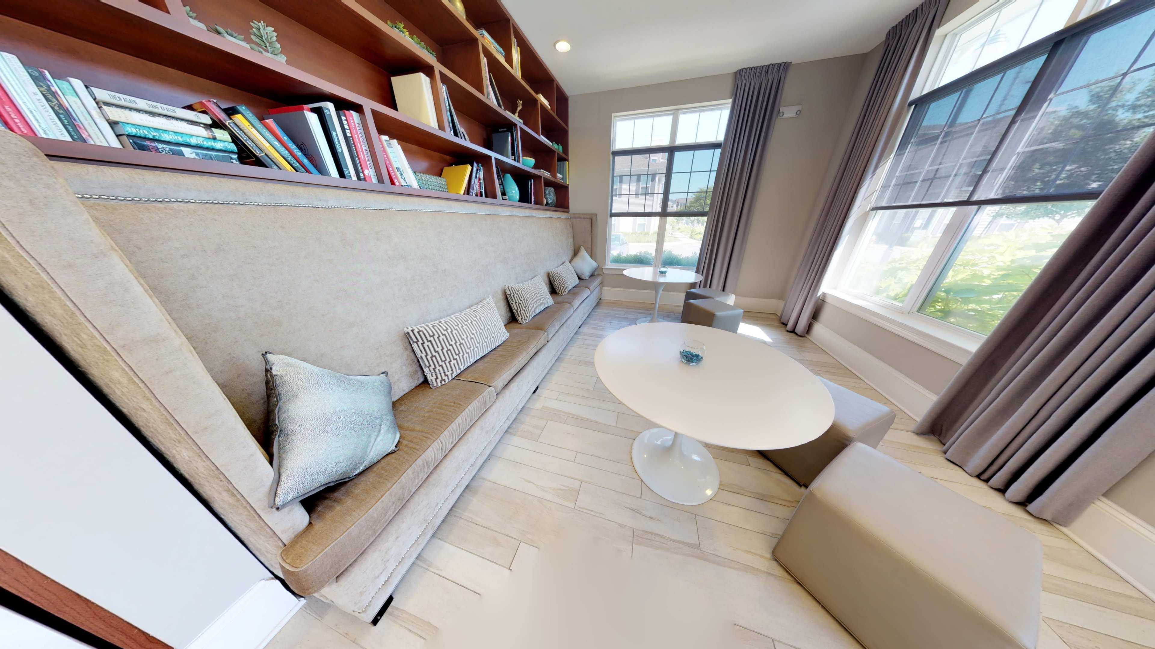A cozy reading nook with a beige sofa, a circular white table, and bookshelves filled with books, illuminated by natural light from a large window.
