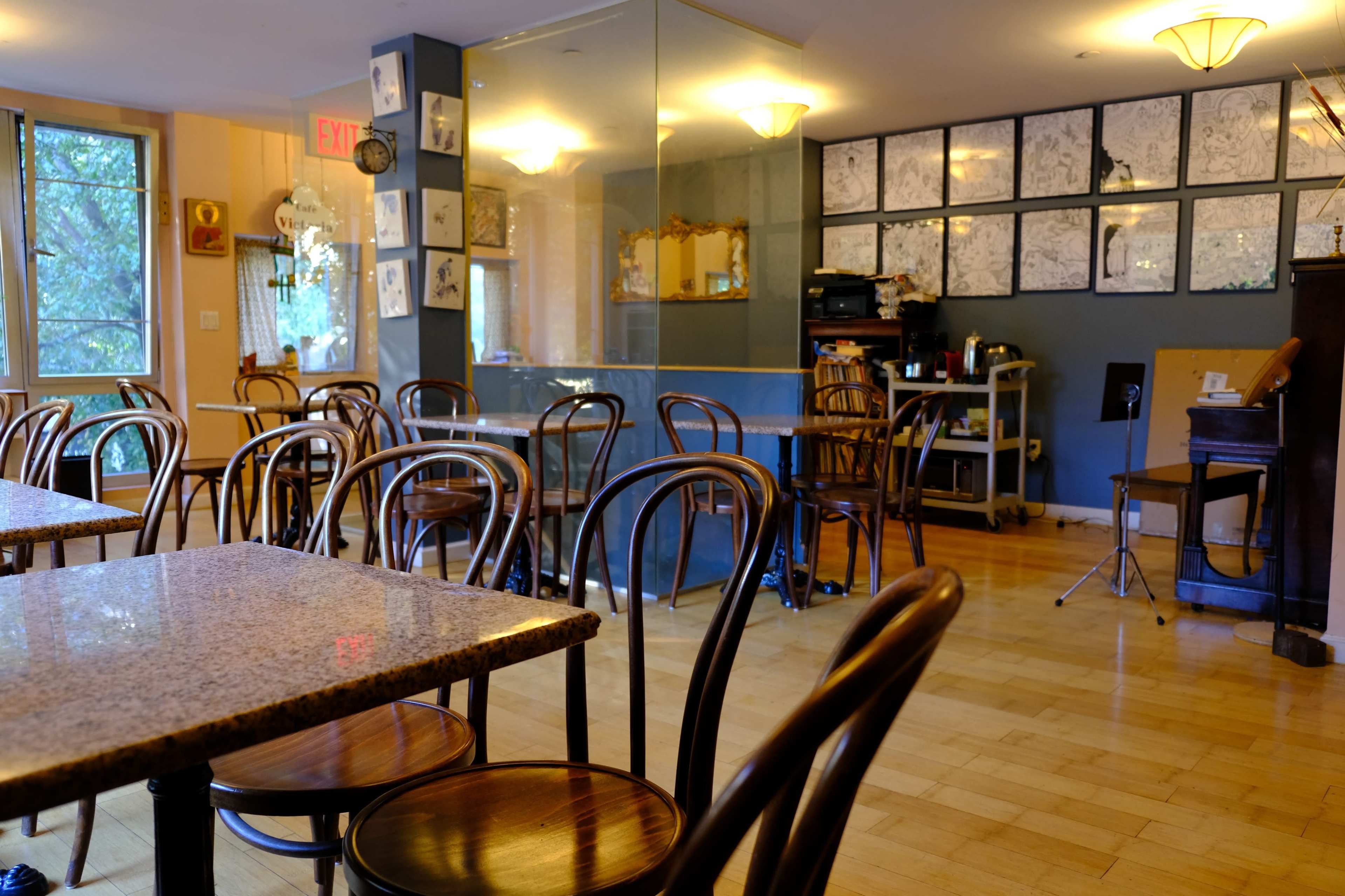 An empty café interior featuring wooden tables and chairs, with a wall adorned with framed sketches and a corner equipped with a piano.