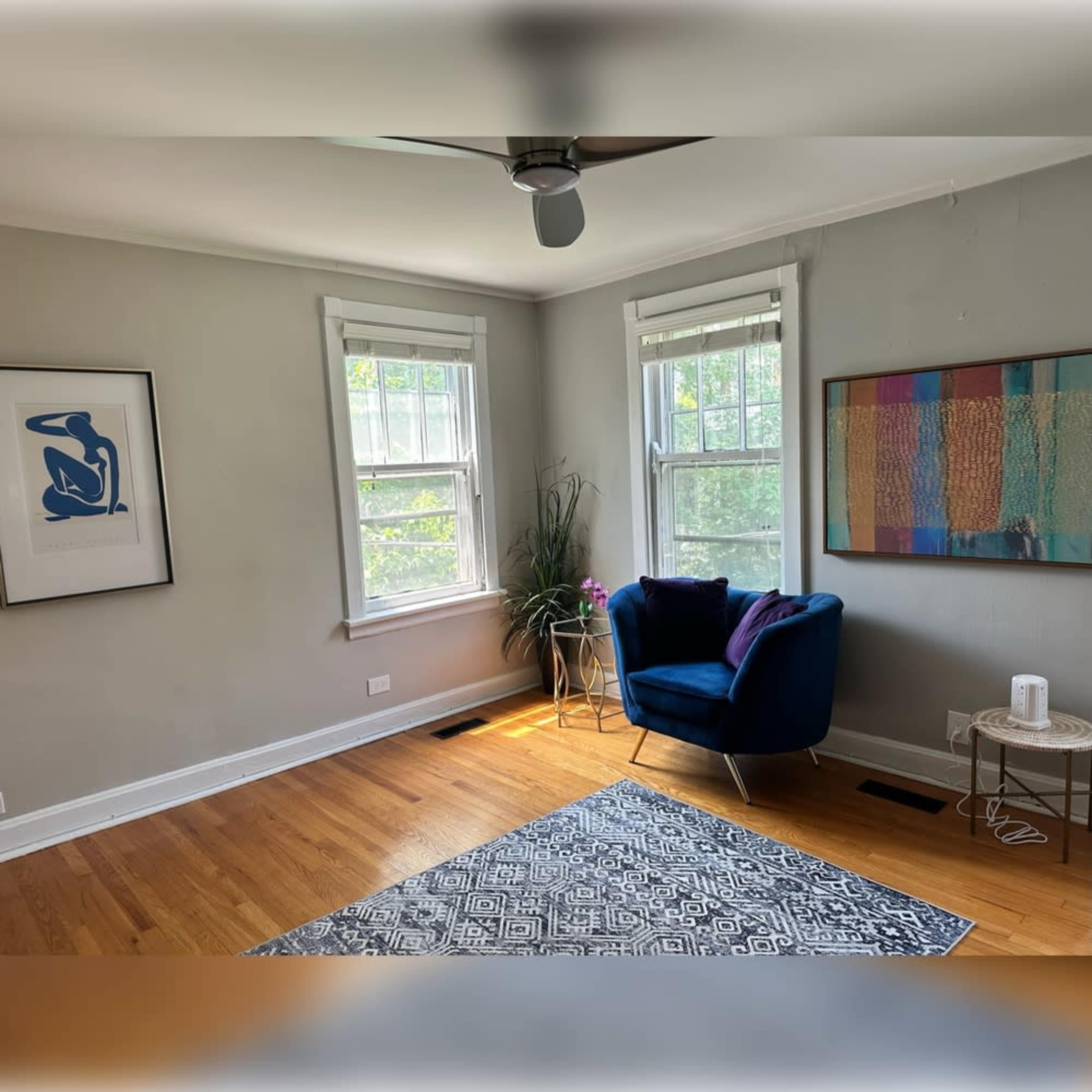 The image shows a well-lit room featuring a blue armchair, a small side table, a rug, and artwork on the walls, along with large windows allowing natural light to enter.
