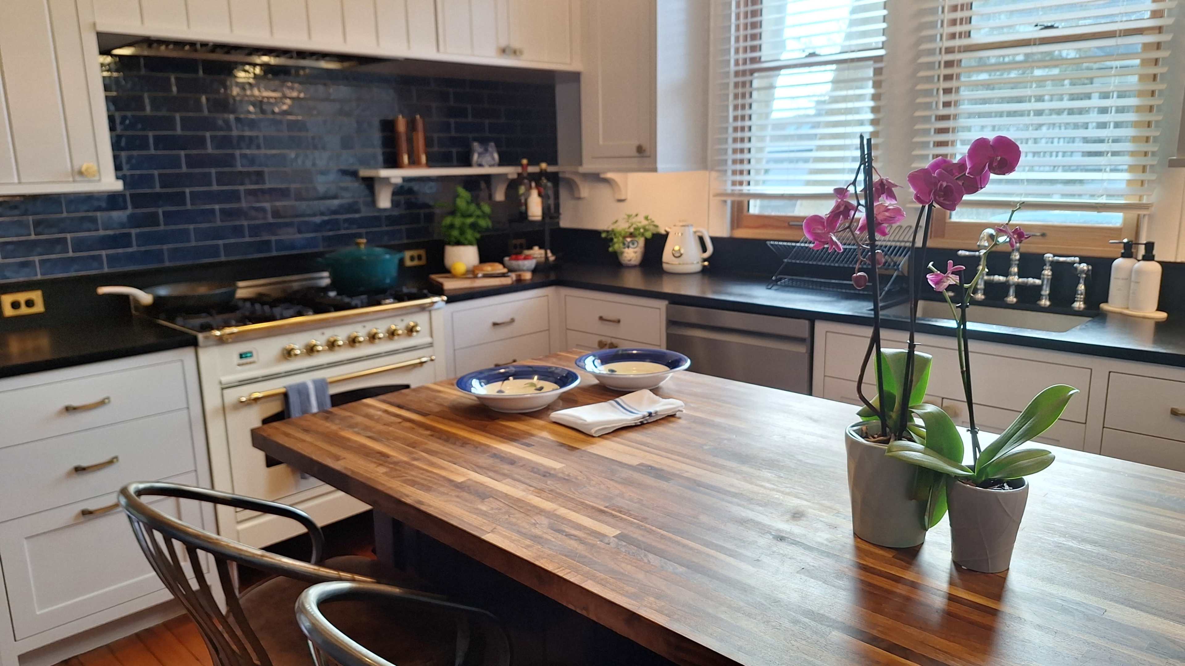 A modern kitchen features a wooden dining table with two bowls, surrounded by white cabinetry, a blue tile backsplash, and plants near a sink.