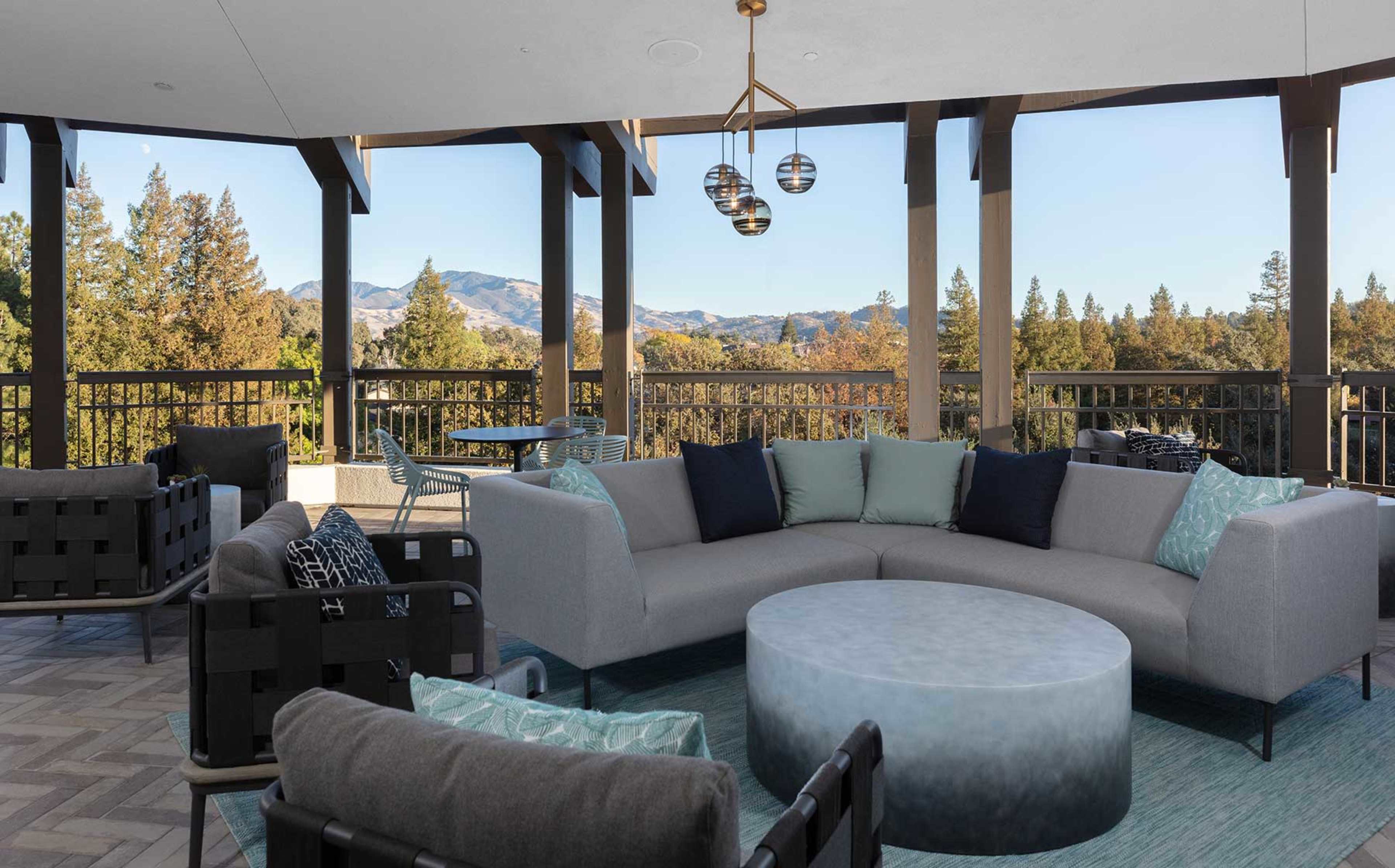The image features a modern outdoor seating area with a large curved sofa, a circular coffee table, and scenic views of rolling hills and trees.