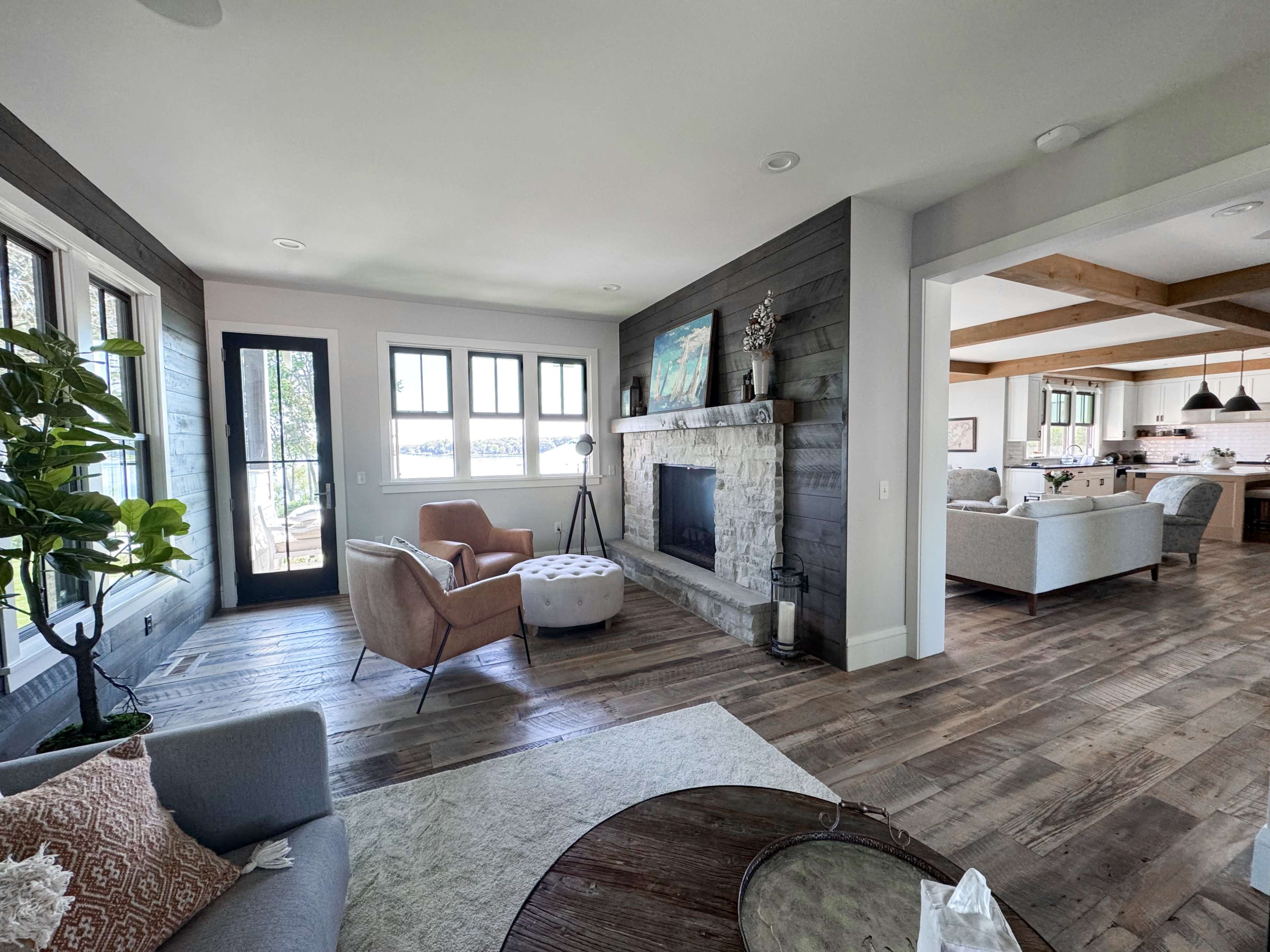 The image shows a living room with a stone fireplace, large windows, and a mix of modern furniture, including two seating areas and a coffee table.
