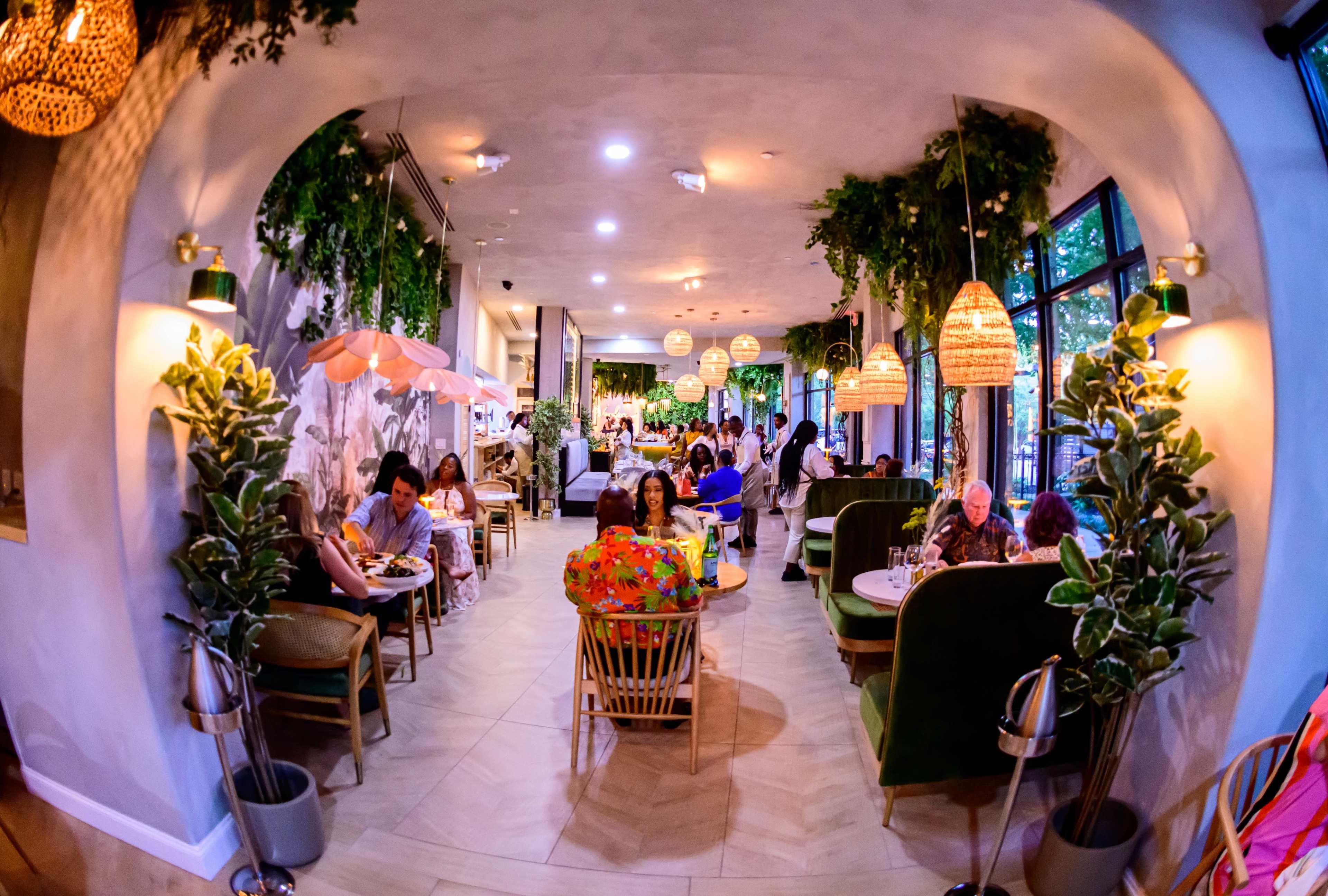 The image shows a vibrant restaurant interior with various patrons seated at tables surrounded by greenery and decorative lighting.