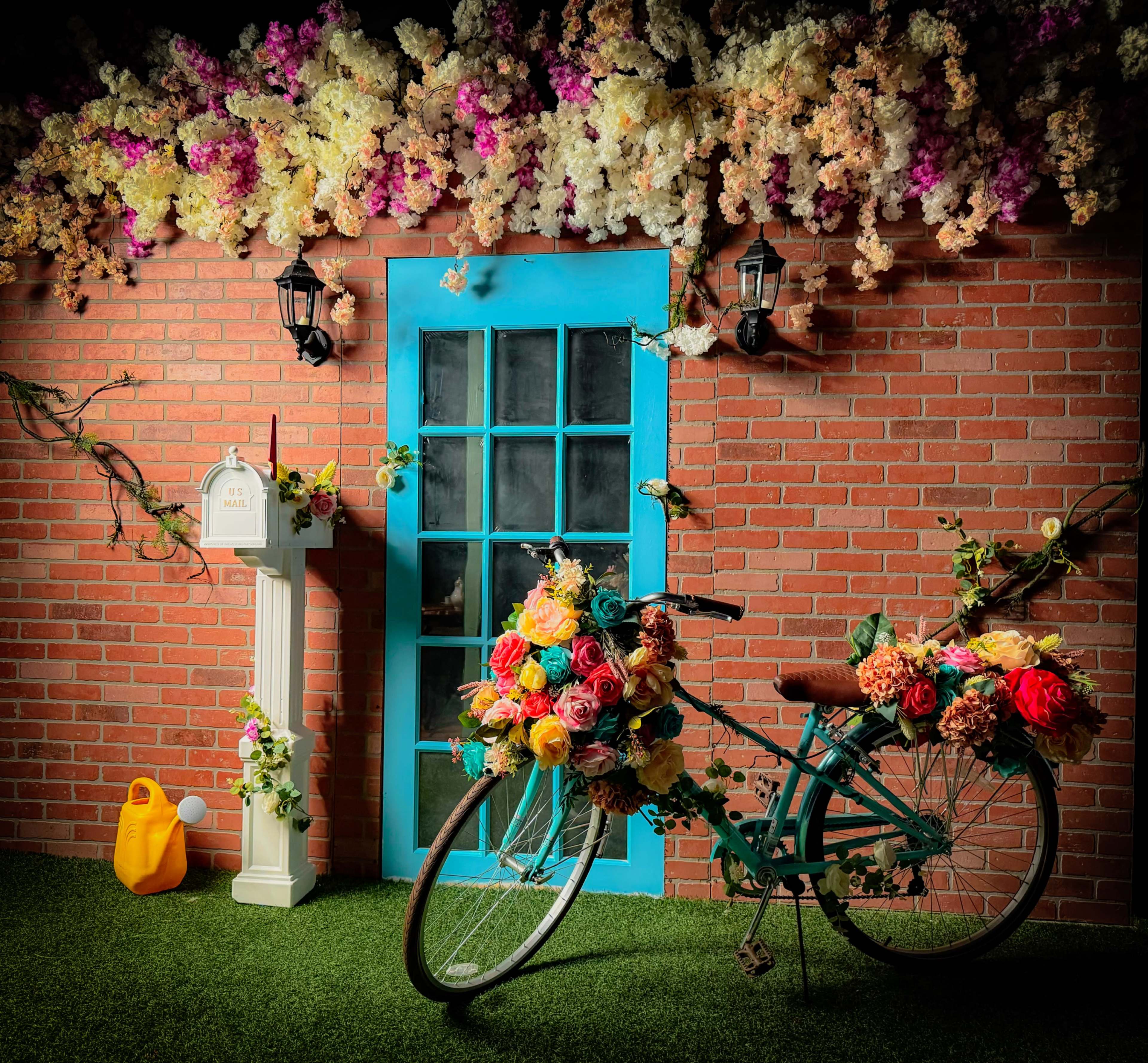 A bicycle adorned with colorful flowers is parked in front of a blue door surrounded by a brick wall and floral decorations.
