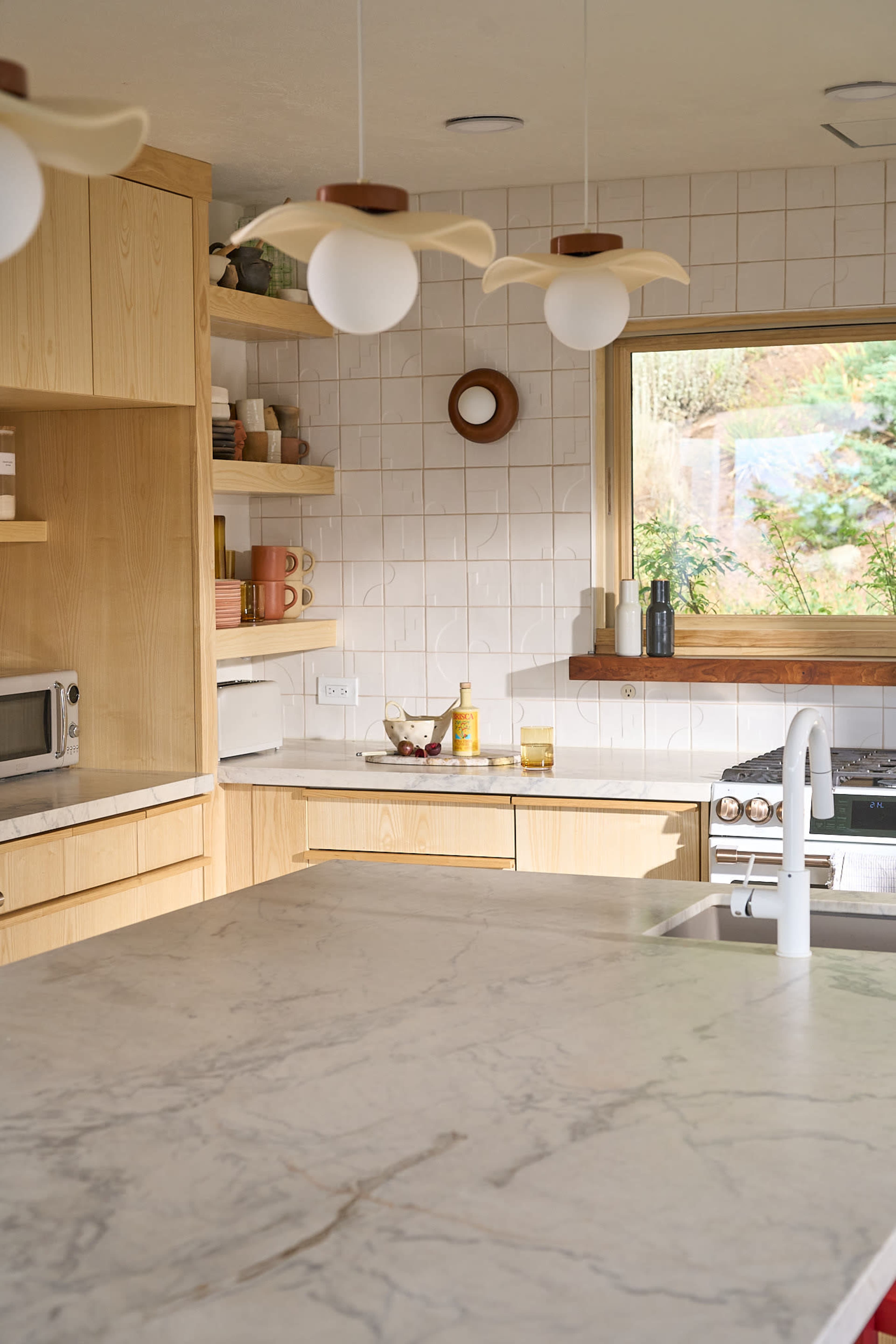 A modern kitchen features wooden cabinetry, marble countertops, and a large window illuminating the space.